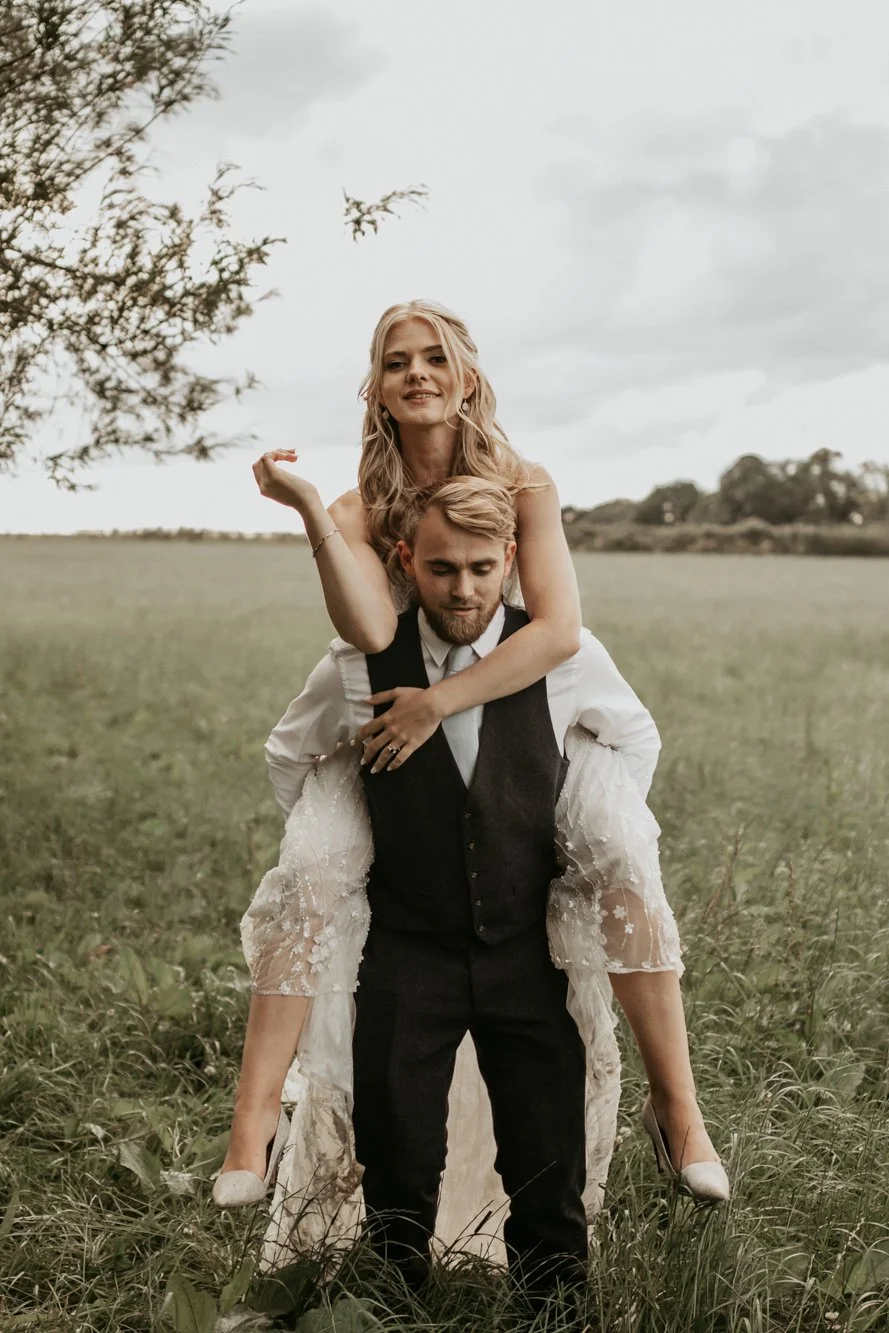 A man in a black vest and white shirt carrying a woman in a wedding dress on his shoulders in a grassy field.