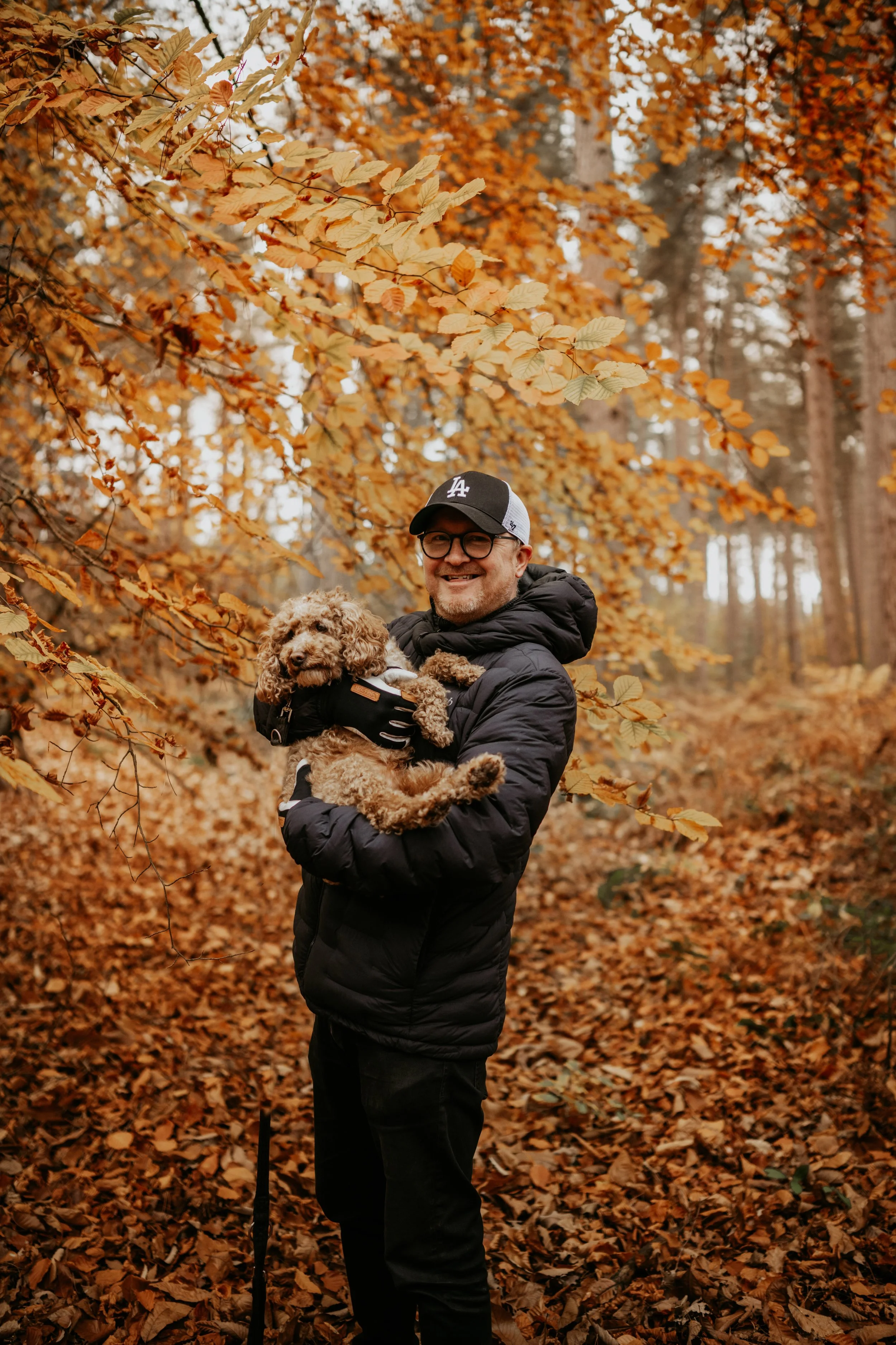 A man in a black jacket, glasses, and a baseball cap holding a small curly-haired dog, standing amidst autumn leaves in a forest.