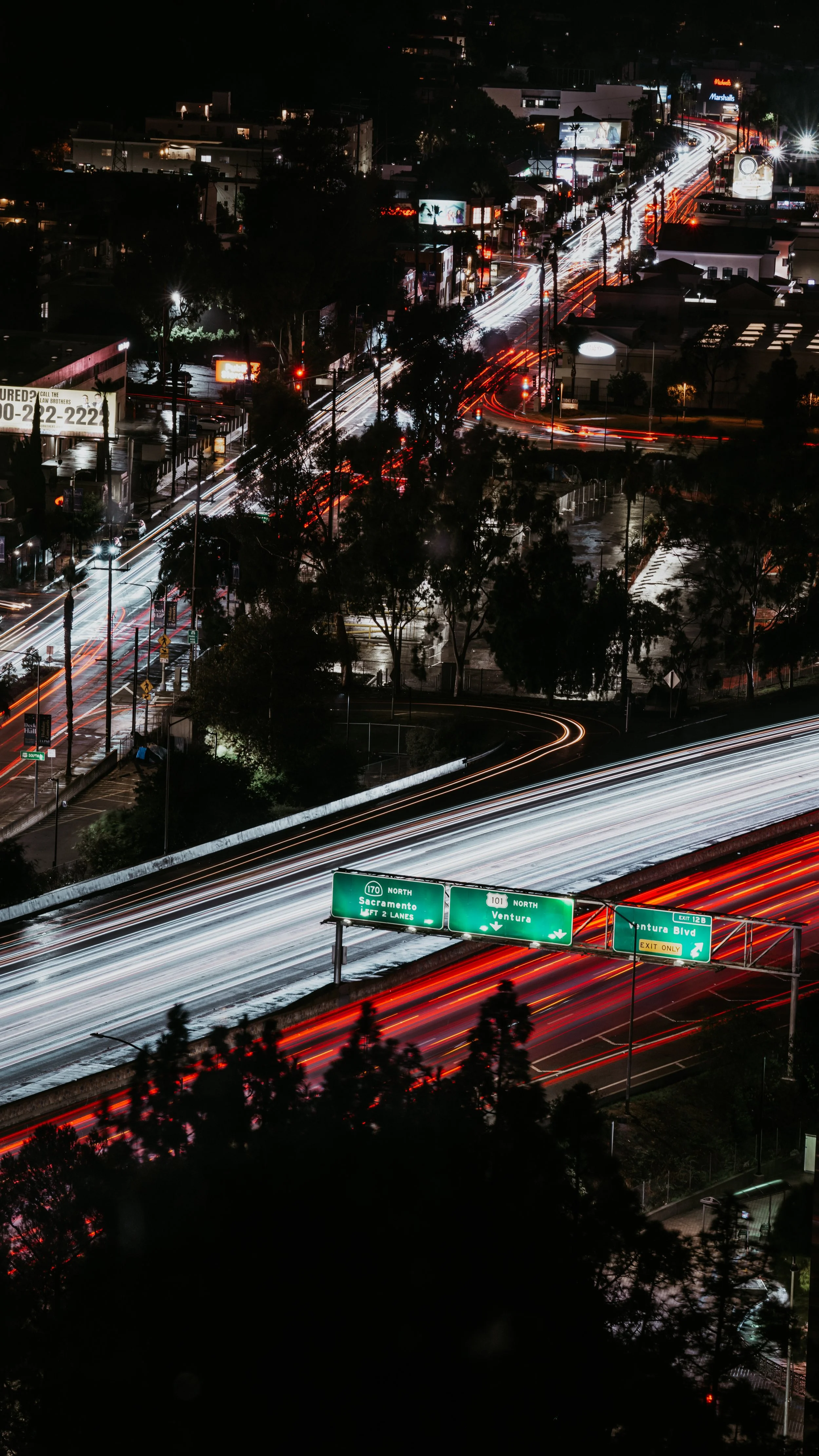 Nighttime cityscape showing busy highways with light trails from moving vehicles, illuminated street signs, and scattered buildings in the background.