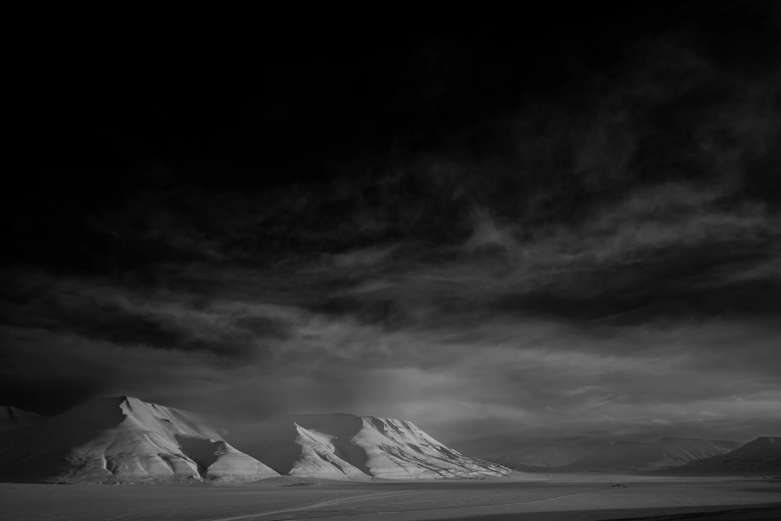 Snow-covered mountains under a dark, cloudy sky in a black and white landscape photograph.