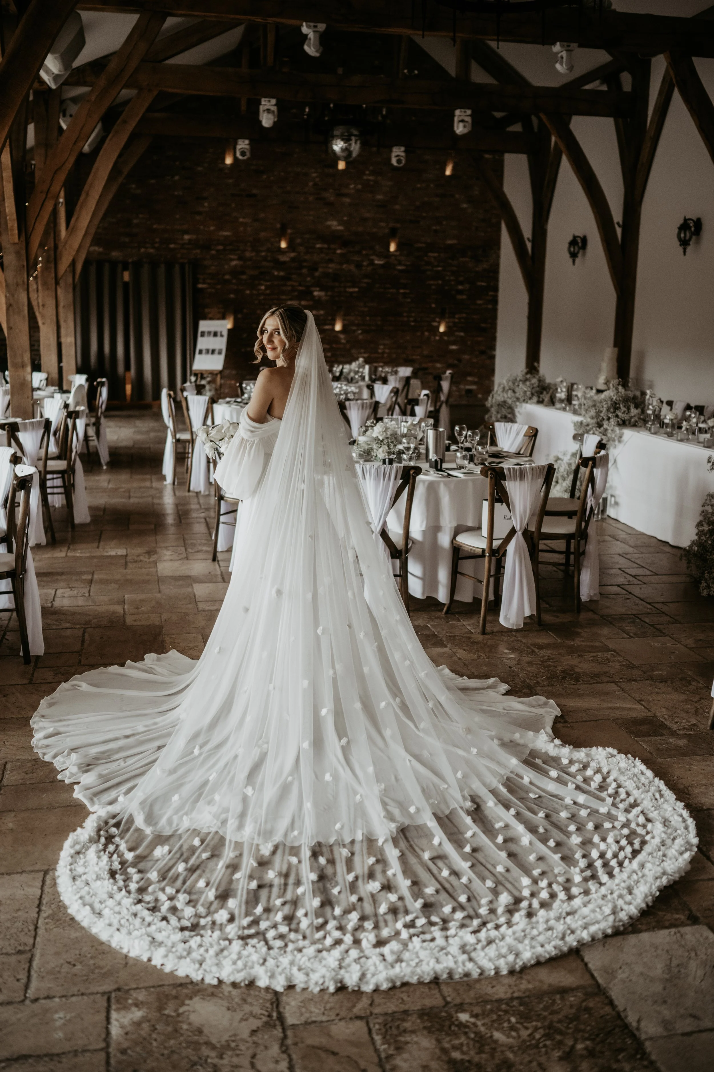 A bride in a white wedding gown with a long veil in a decorated reception hall, surrounded by round tables with white tablecloths and chairs.