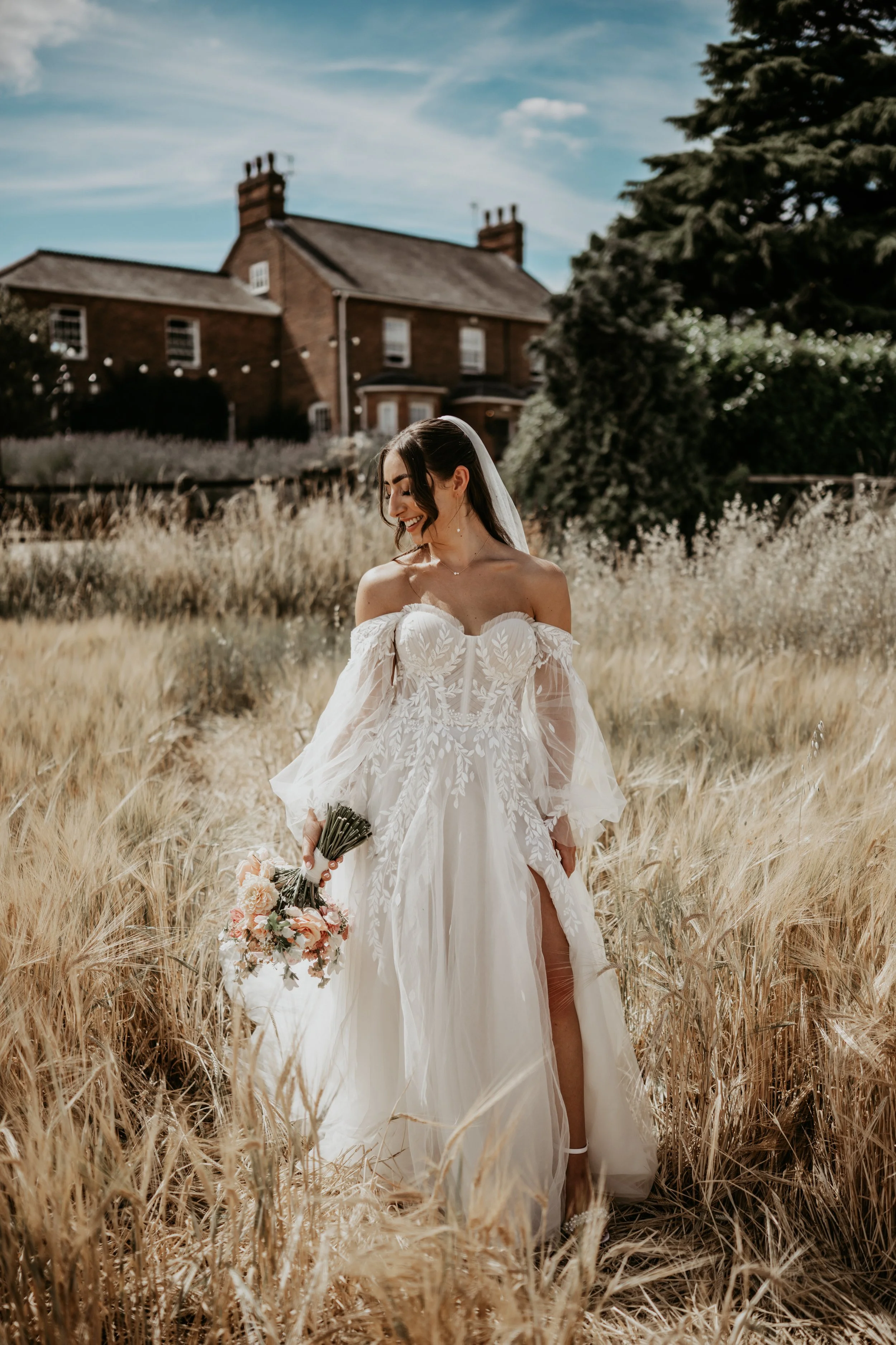 A bride in a white wedding dress with off-shoulder sleeves standing in a field of tall, golden grass, holding a bouquet, smiling, with a brick house and trees in the background on a sunny day.