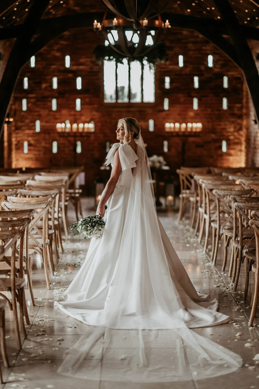 A bride in a white wedding dress standing in an elegant chapel with wooden walls and rows of chairs, holding a bouquet of flowers, with candlelit sconces and candles on the wall behind her.