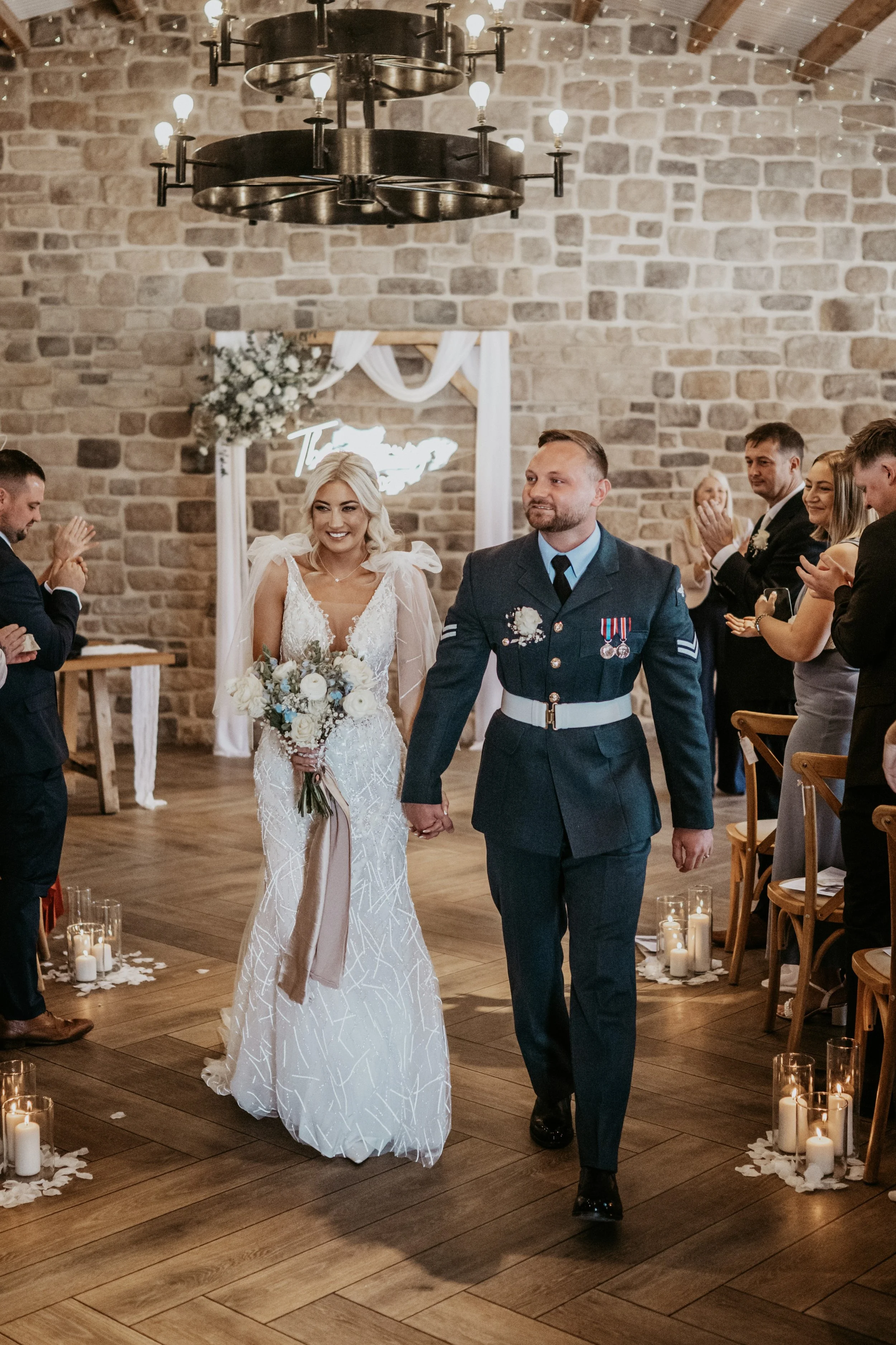 A bride and groom walking hand in hand down the aisle at their wedding in a decorated indoor venue with candles and guests clapping.