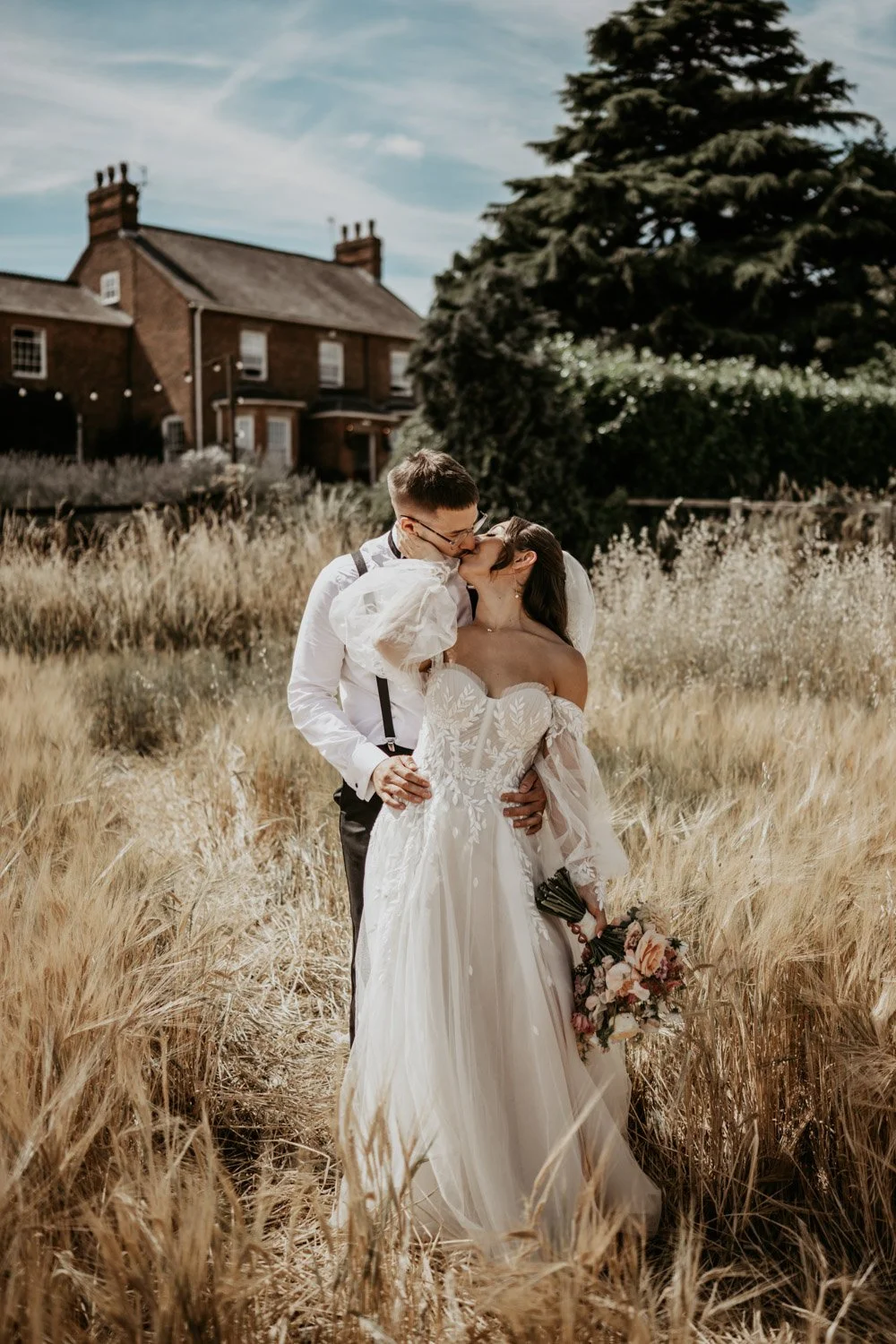 A newlywed couple sharing a kiss in a field of tall, golden grass, with a large house and trees in the background on a sunny day.