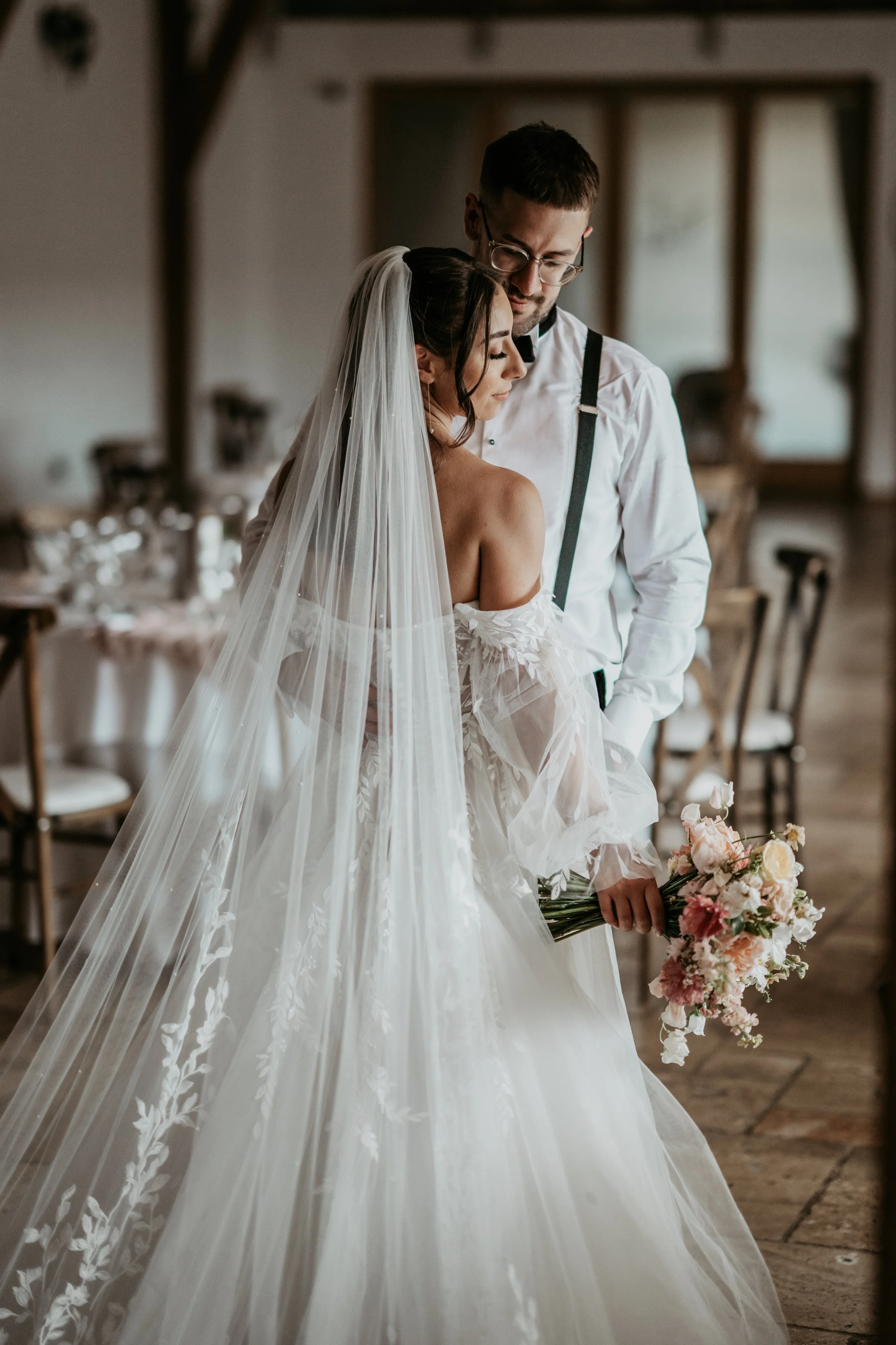 Bride in a white wedding dress with veil holding a bouquet, standing closely with a groom in a white shirt and suspenders, inside a rustic wooden venue.