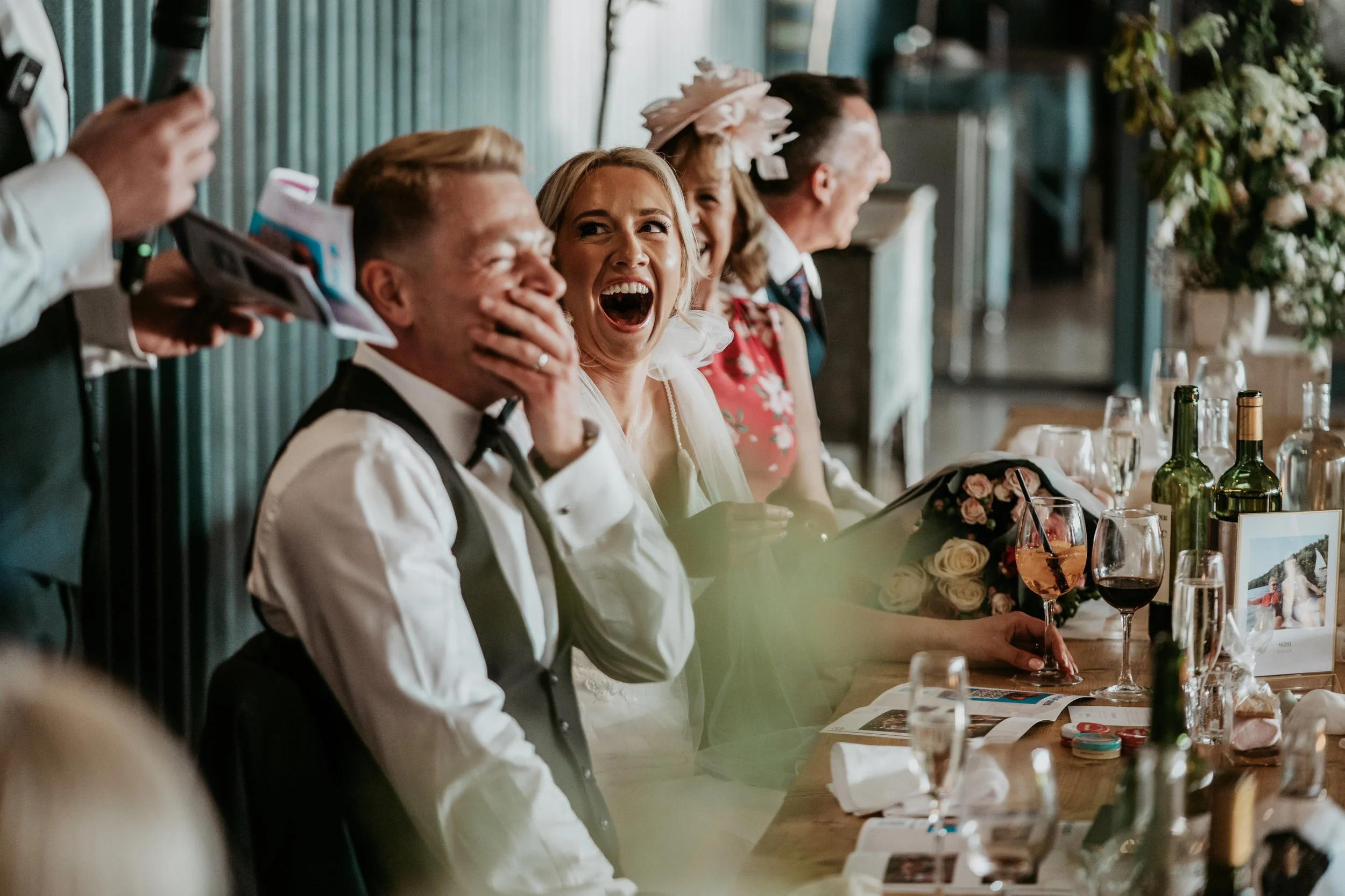 People sitting at a decorated table during a celebration, with some laughing and others smiling, glasses of wine and flowers on the table.