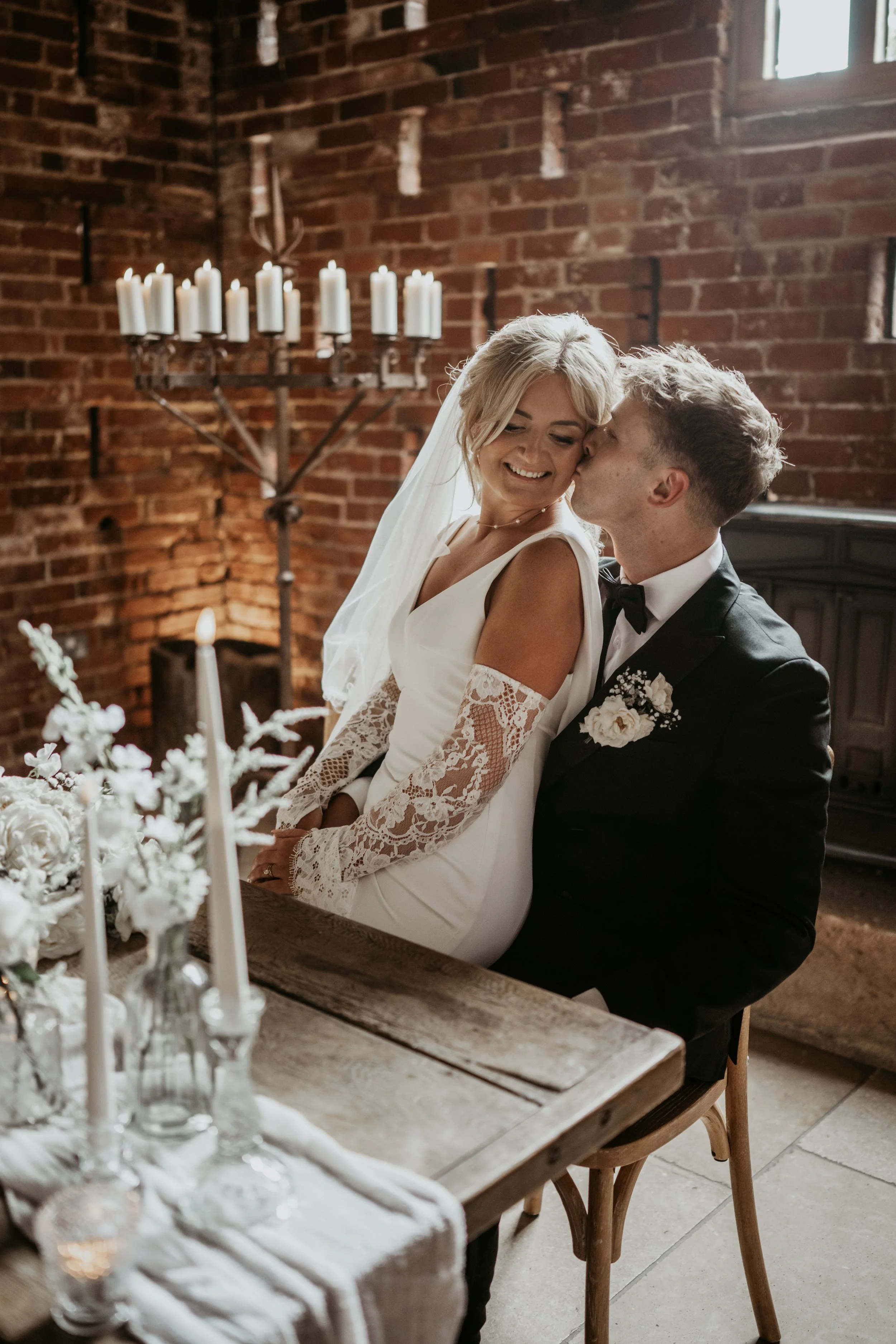 A bride and groom enjoying a moment together during their wedding in a rustic indoor setting with brick walls, candles, and floral decorations.