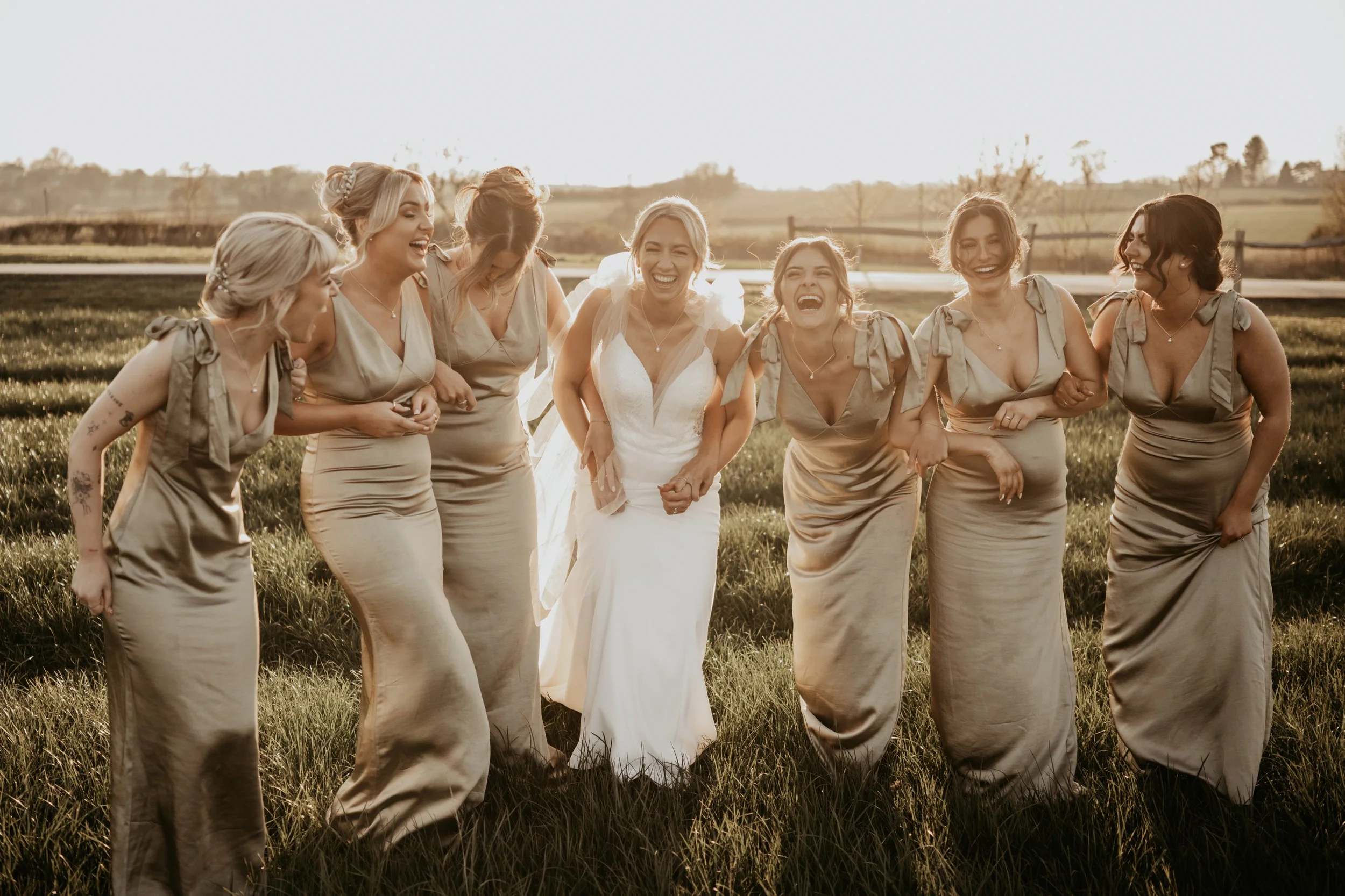 A group of women, including a bride in a white dress, laughing and walking together in a field at sunset.