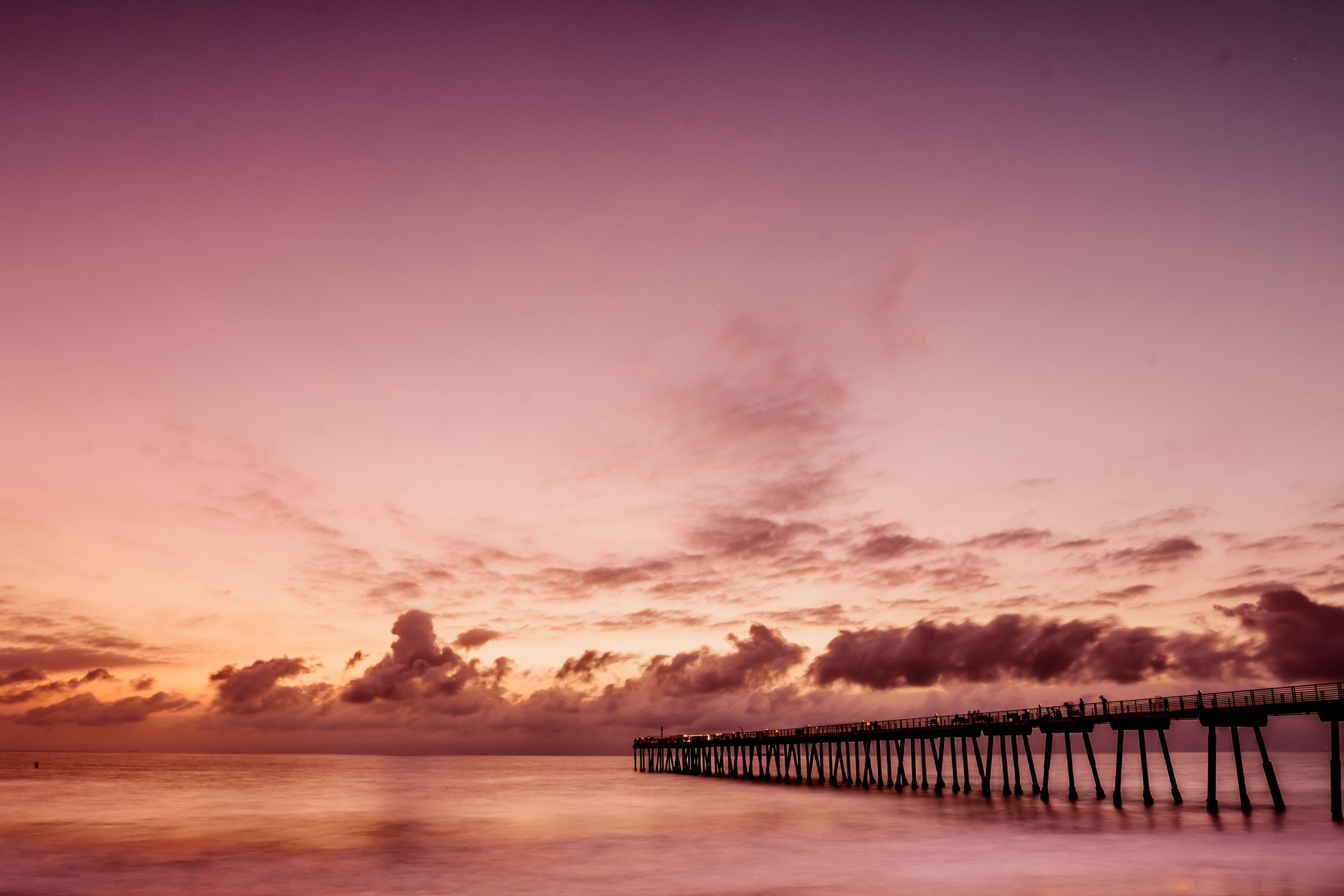 Sunset view over the ocean with a pier extending into the water, pink and purple sky with clouds.