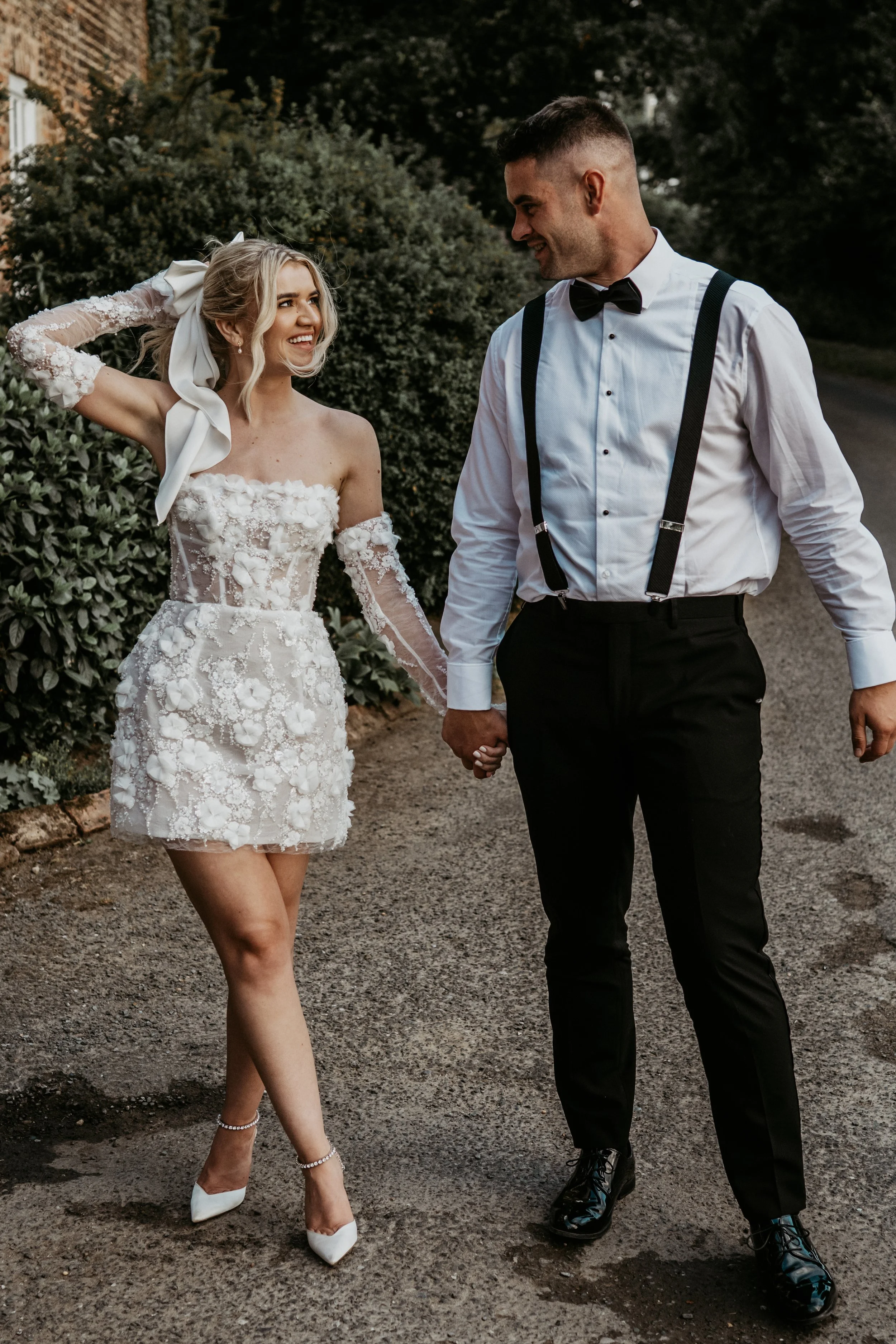 A smiling bride in a lace wedding dress holding hands with a smiling groom in a white shirt, black suspenders, and bow tie, walking outdoors on a gravel path.