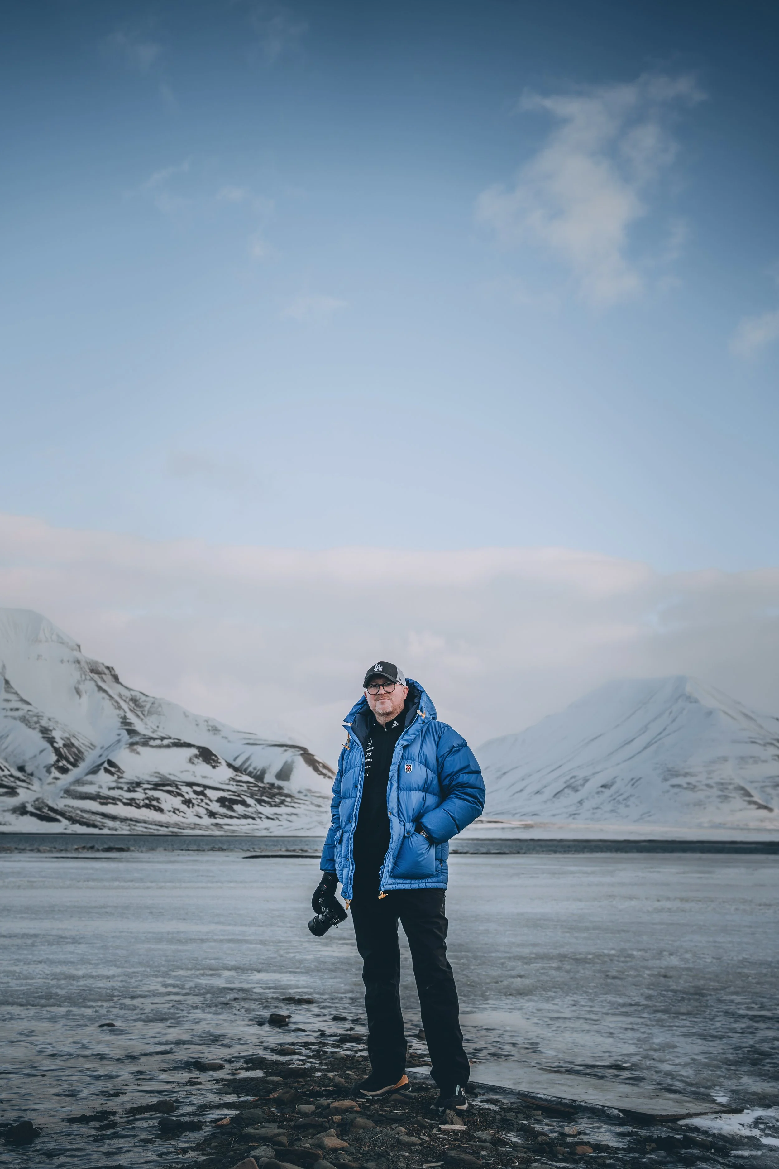 A man standing on a rocky shoreline in a cold, snowy landscape with mountains in the background, dressed in a blue winter jacket and holding a camera.