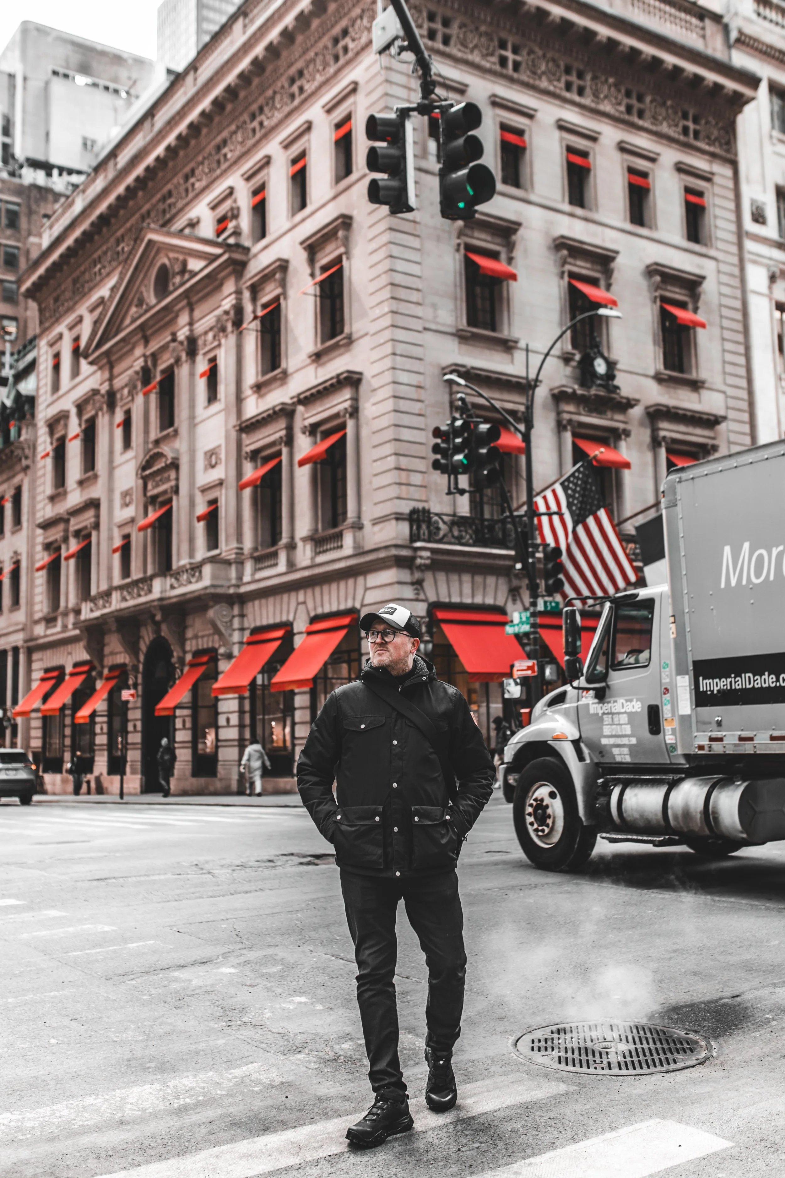 A man walking across a city street with tall buildings, traffic lights, American flags, and a delivery truck in the background.