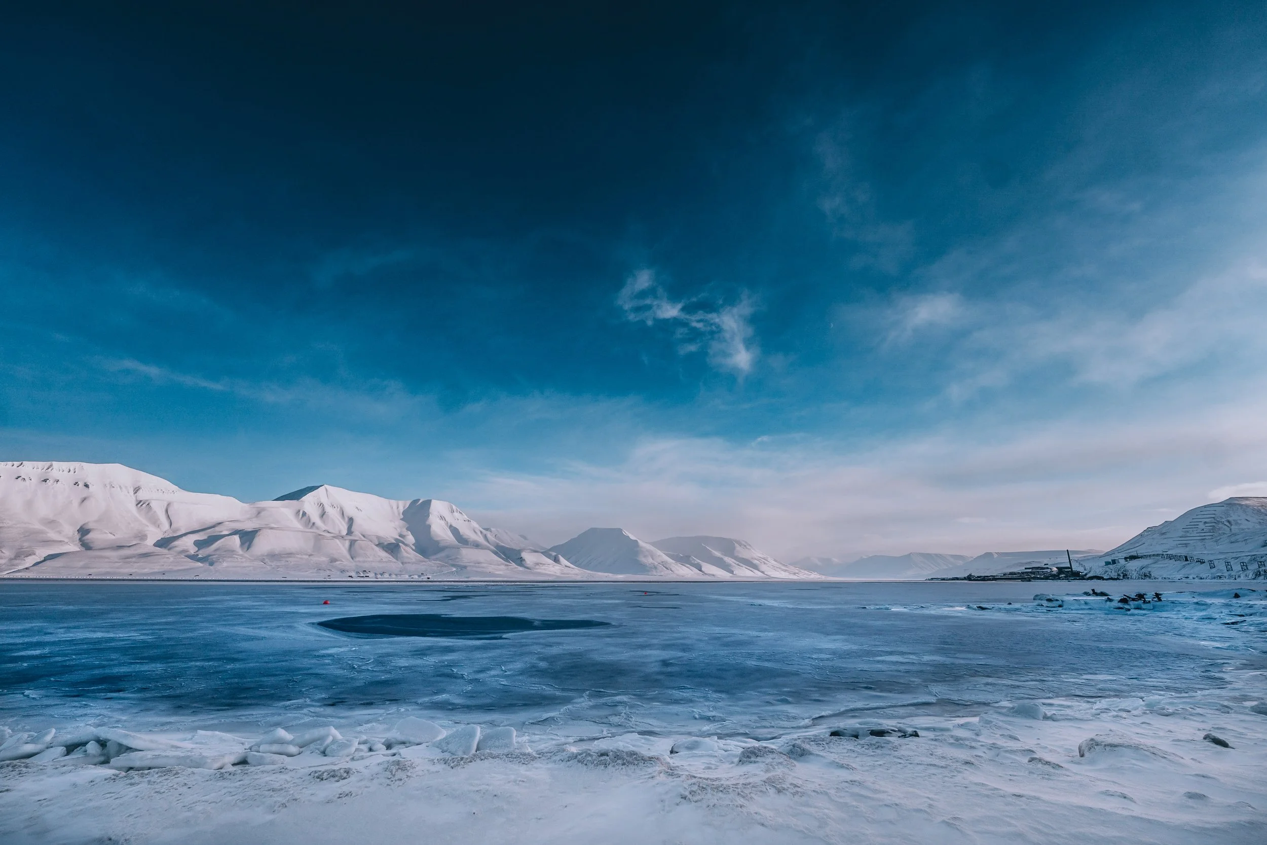 A vast icy landscape featuring snow-covered mountains under a blue sky with some clouds, a frozen sea or lake with open water patches, and a small boat or structure in the distance.