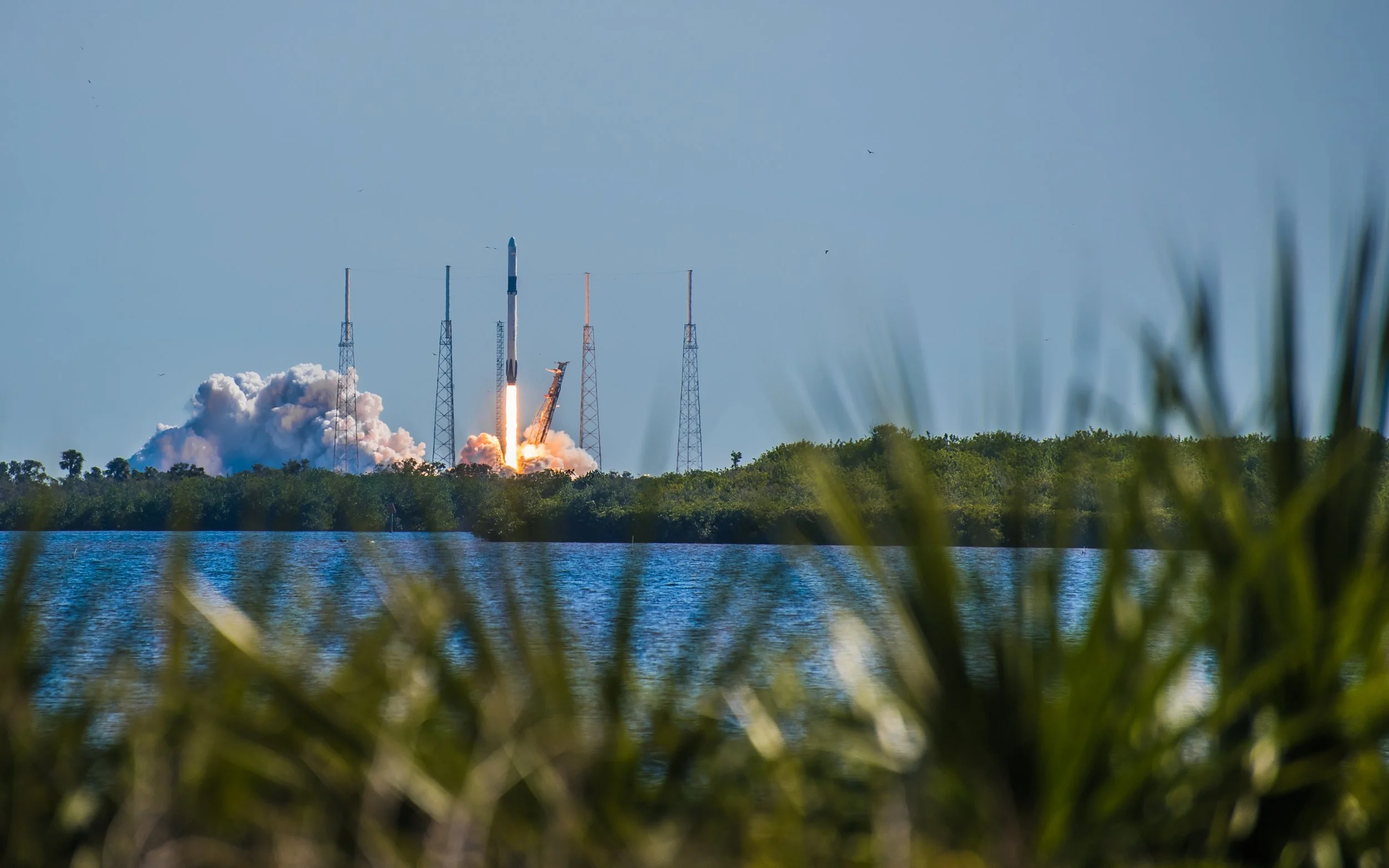 A rocket launching from a space center with a trail of fire and smoke, surrounded by tall communication towers and greenery, over a body of water.