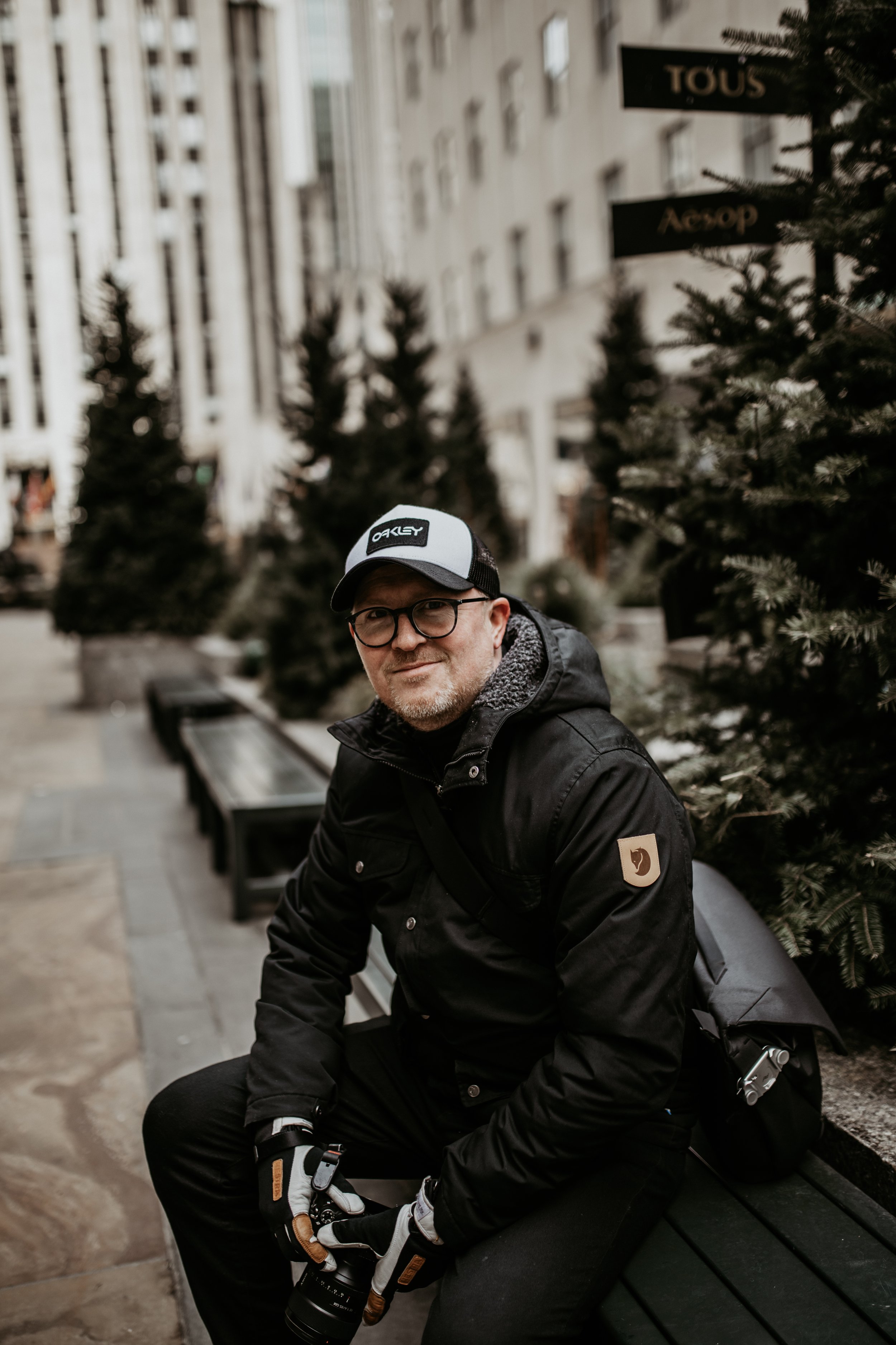 A man sitting on a park bench outdoors, wearing glasses, a black jacket with a patch on the sleeve, a black and white baseball cap, gloves, and holding a camera. There are decorated Christmas trees near him, with buildings and signs in the background.