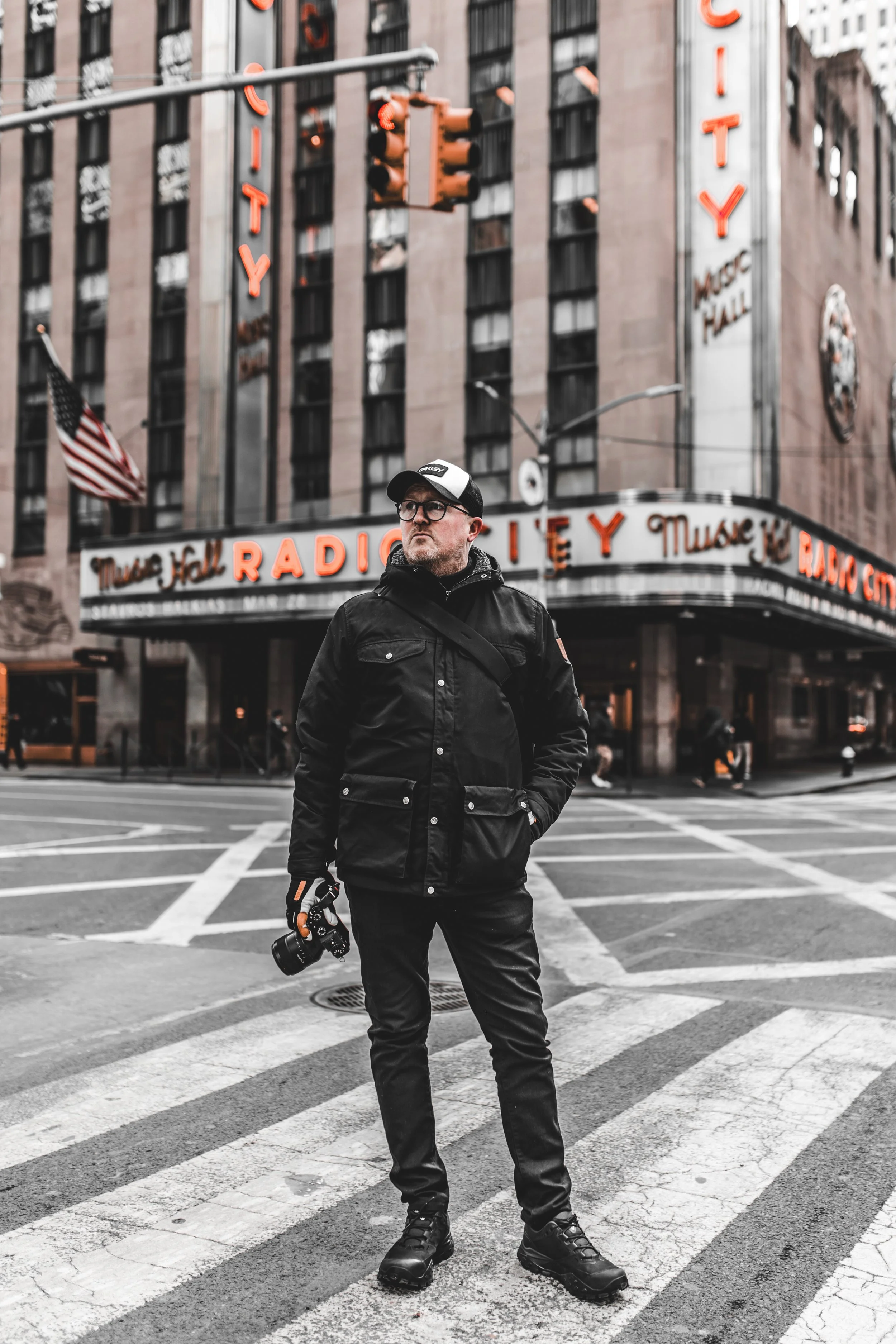 A man standing at a crosswalk on a city street, holding a camera in his right hand, dressed in black, with a radio city music hall sign in the background.