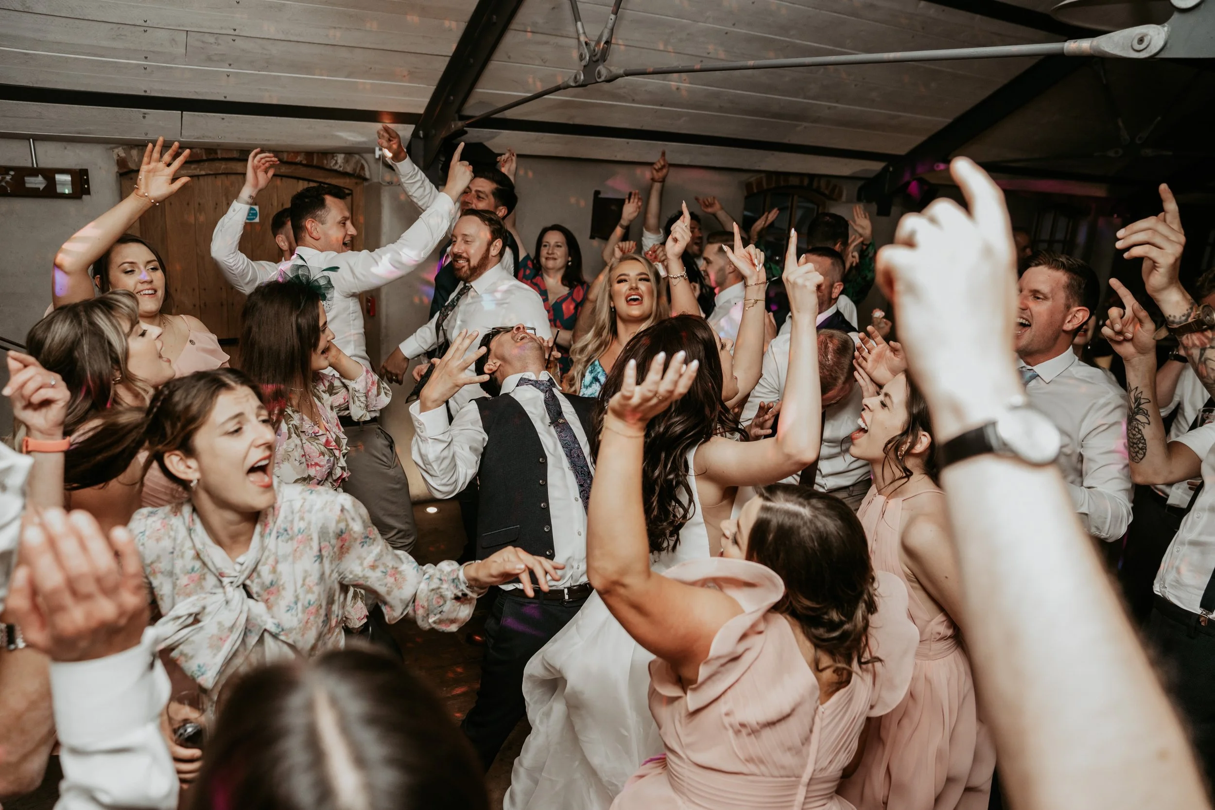 Crowd of people dancing and celebrating at a party or wedding reception, with many raising their hands and smiling.