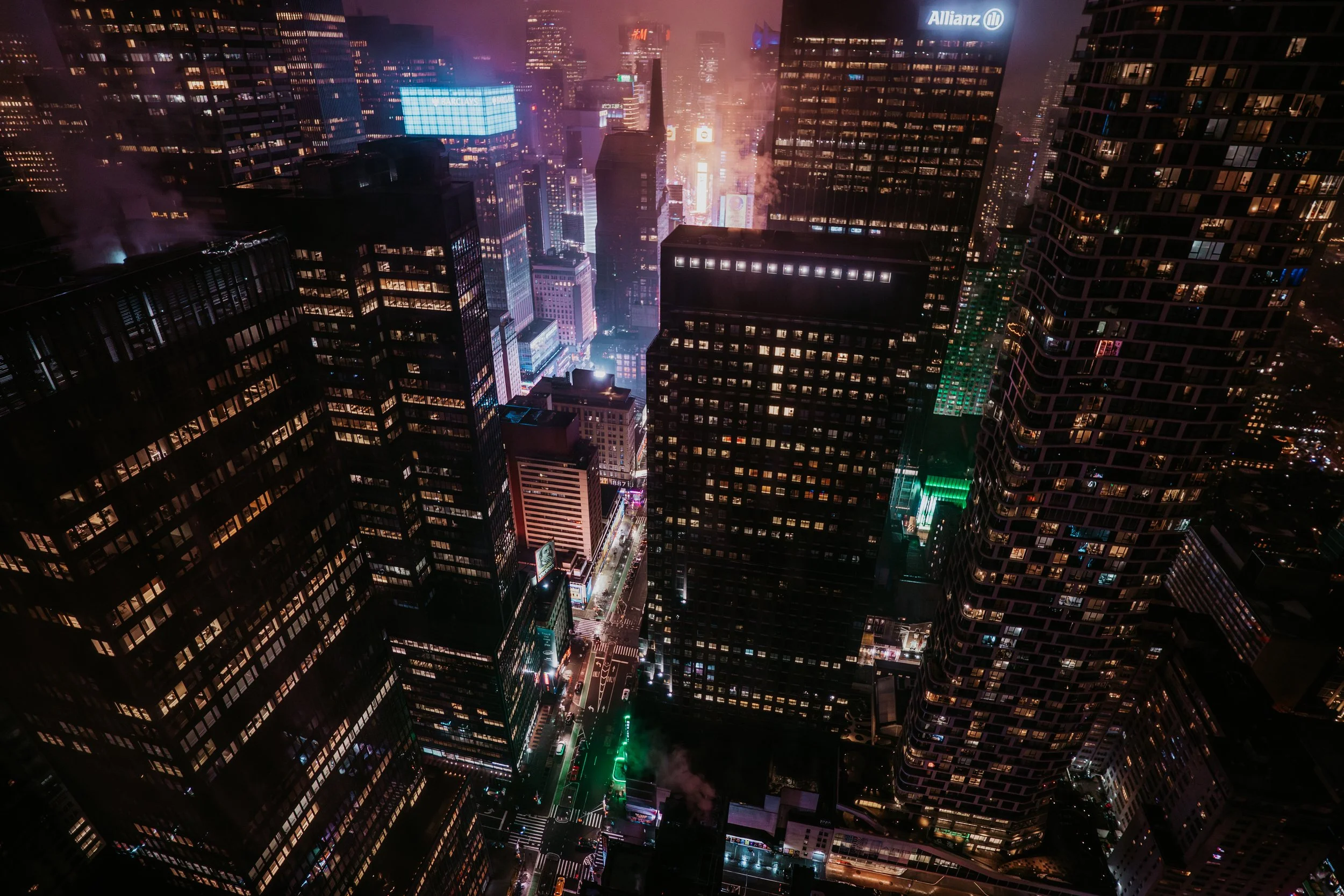 Nighttime aerial view of a city with tall illuminated skyscrapers, glowing neon signs, and streets below, with some buildings emitting smoke or steam.