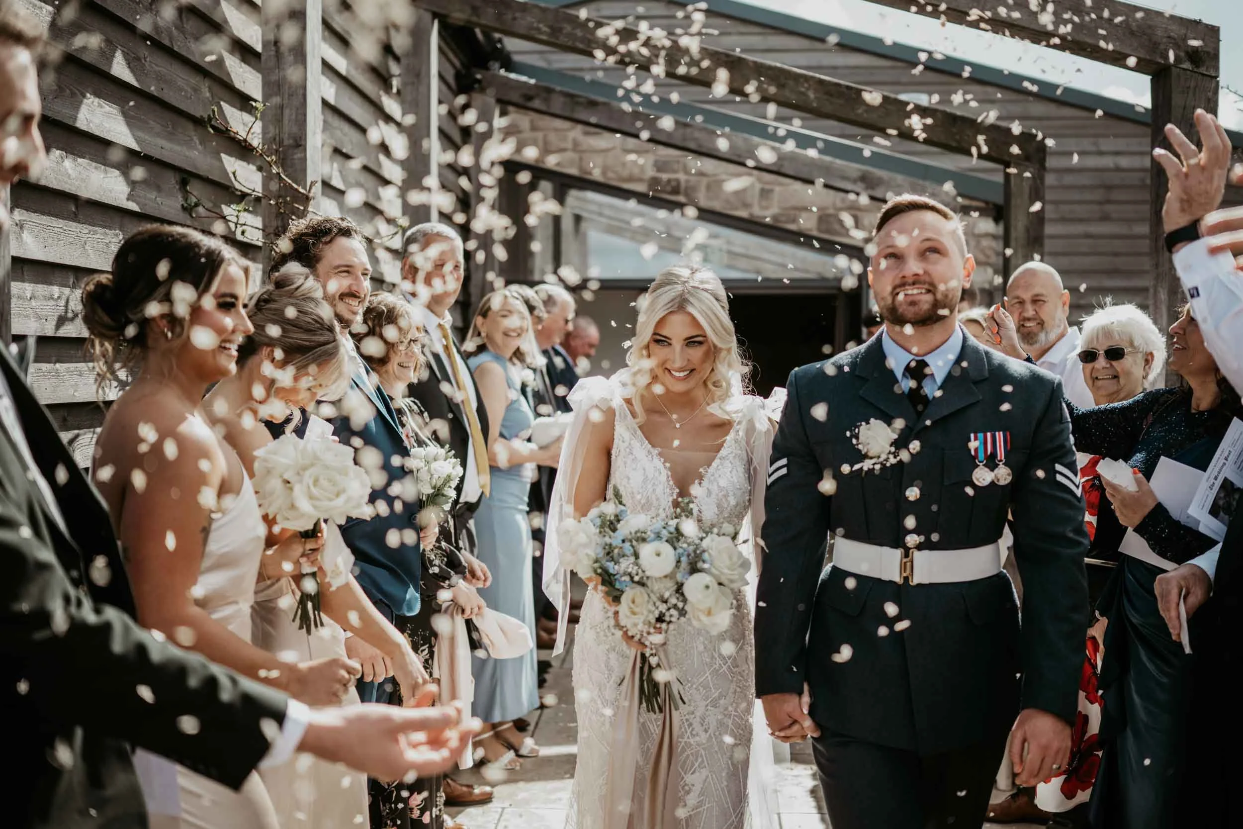 A bride and groom walk hand-in-hand surrounded by wedding guests throwing confetti at an outdoor wedding venue.