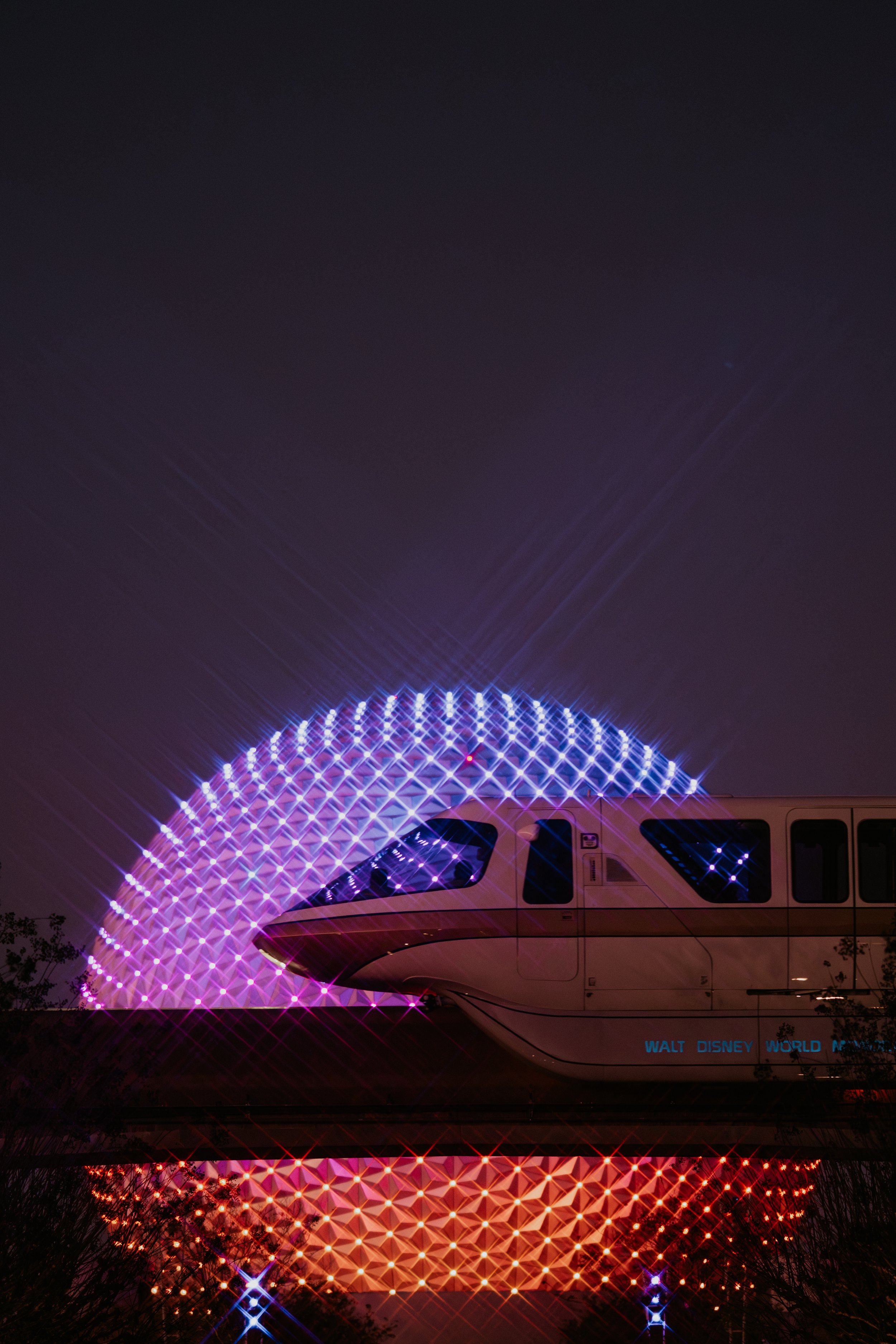 Nighttime view of the Monorail at EPCOT with illuminated Geodesic Sphere (Spaceship Earth) in the background, reflecting on a nearby water surface.