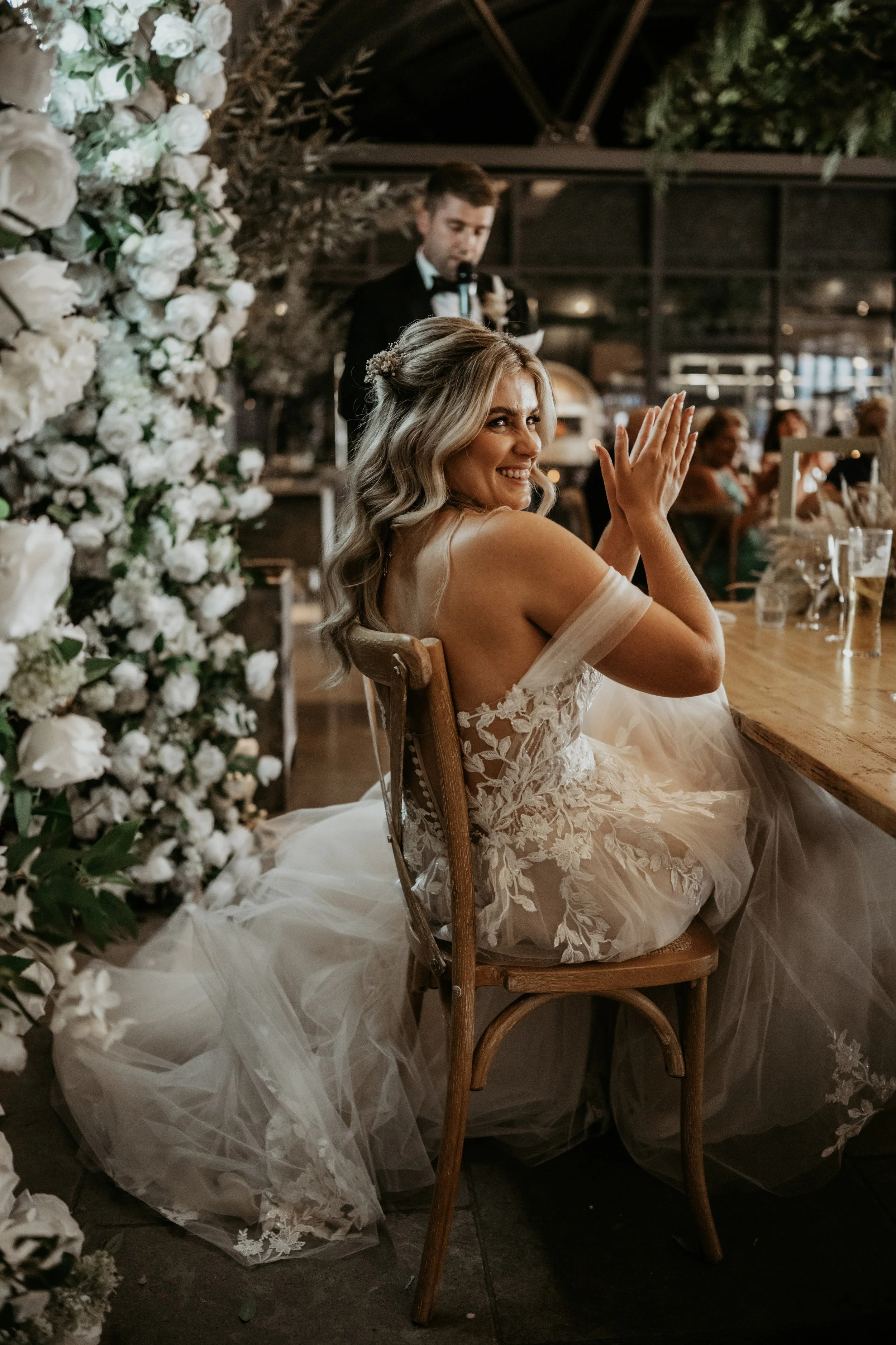 A bride in a wedding dress claps and smiles as she sits at a decorated table during her wedding reception, with a floral backdrop and a man speaking in the background.