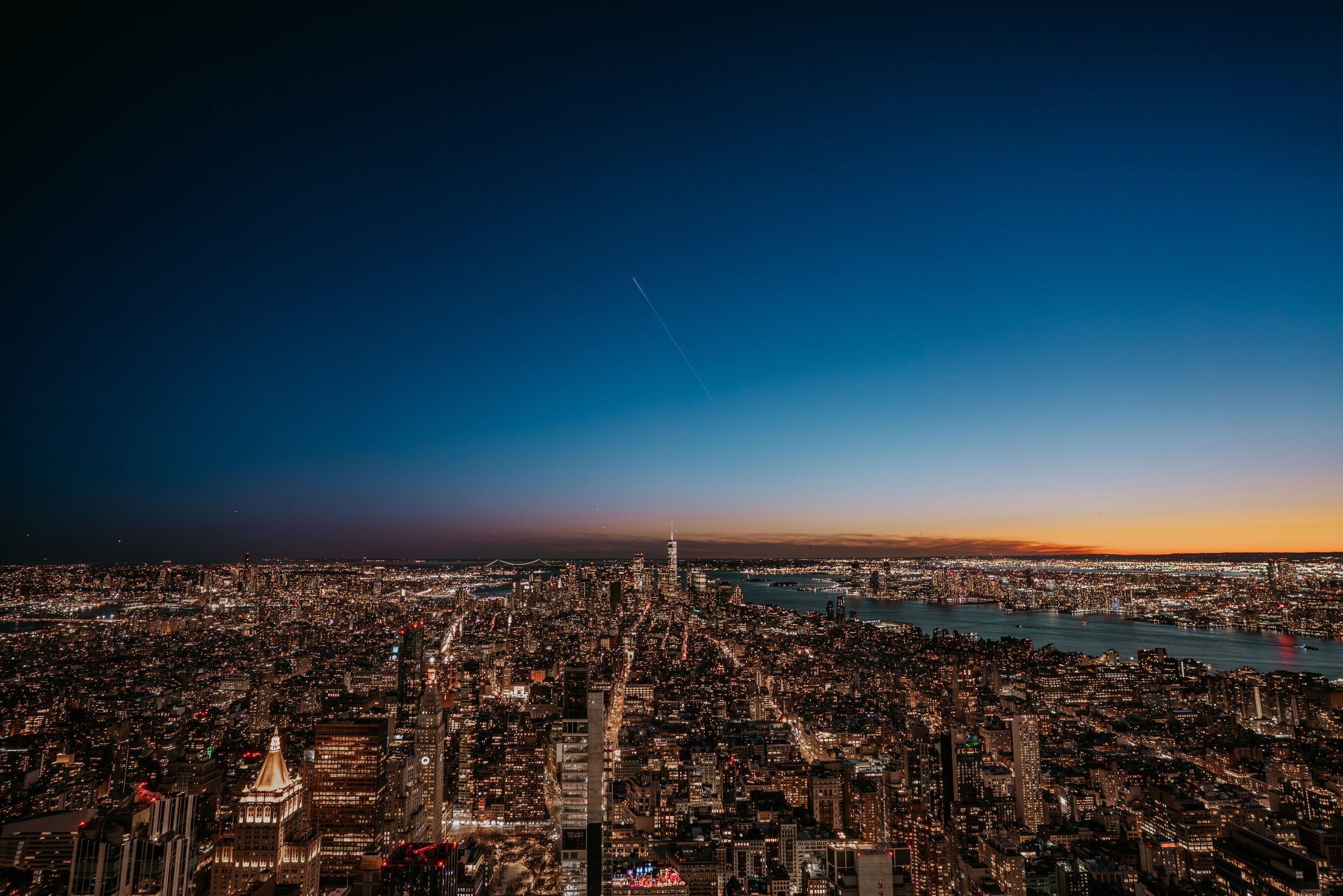 An aerial view of New York City skyline at dusk, featuring tall skyscrapers, including the Empire State Building, with a river and bridges in the background, under a clear evening sky.