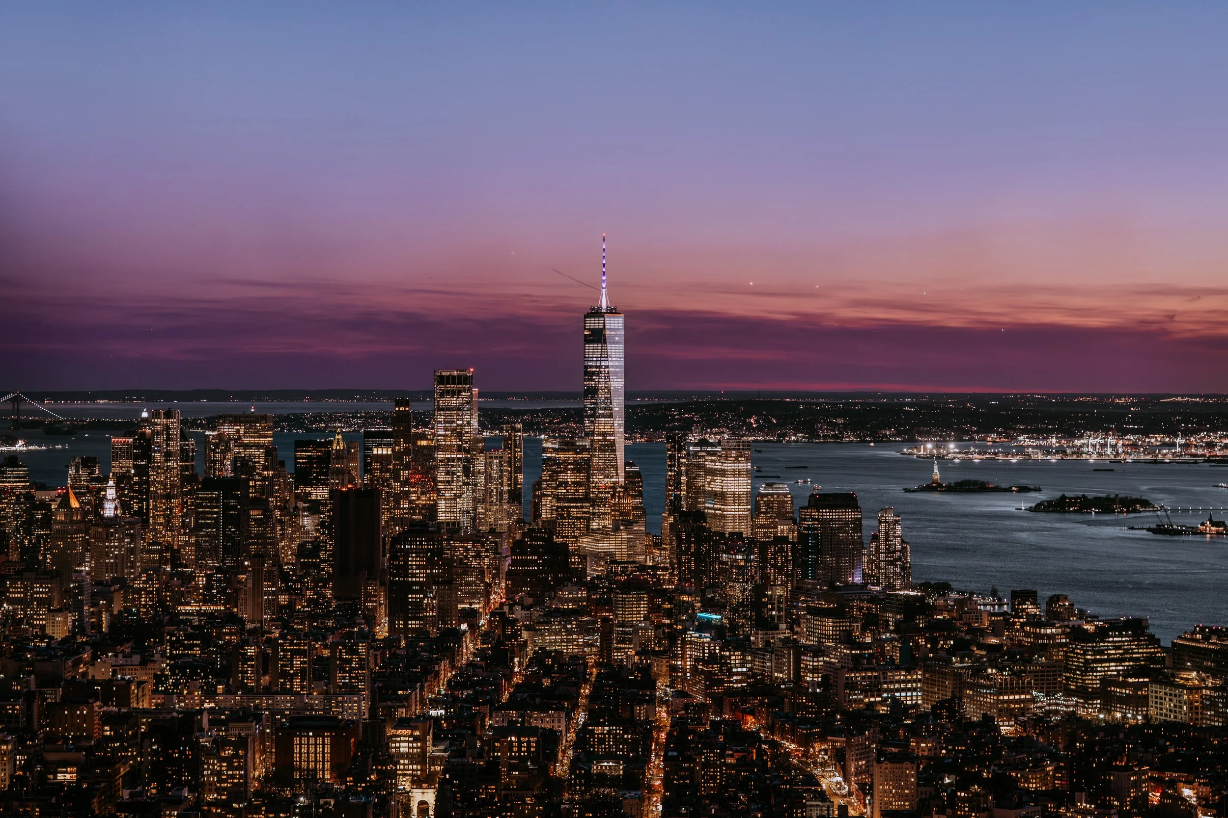 Sunset over Midtown Manhattan skyline with One World Trade Center illuminated, city lights, Hudson River, and distant bridges.