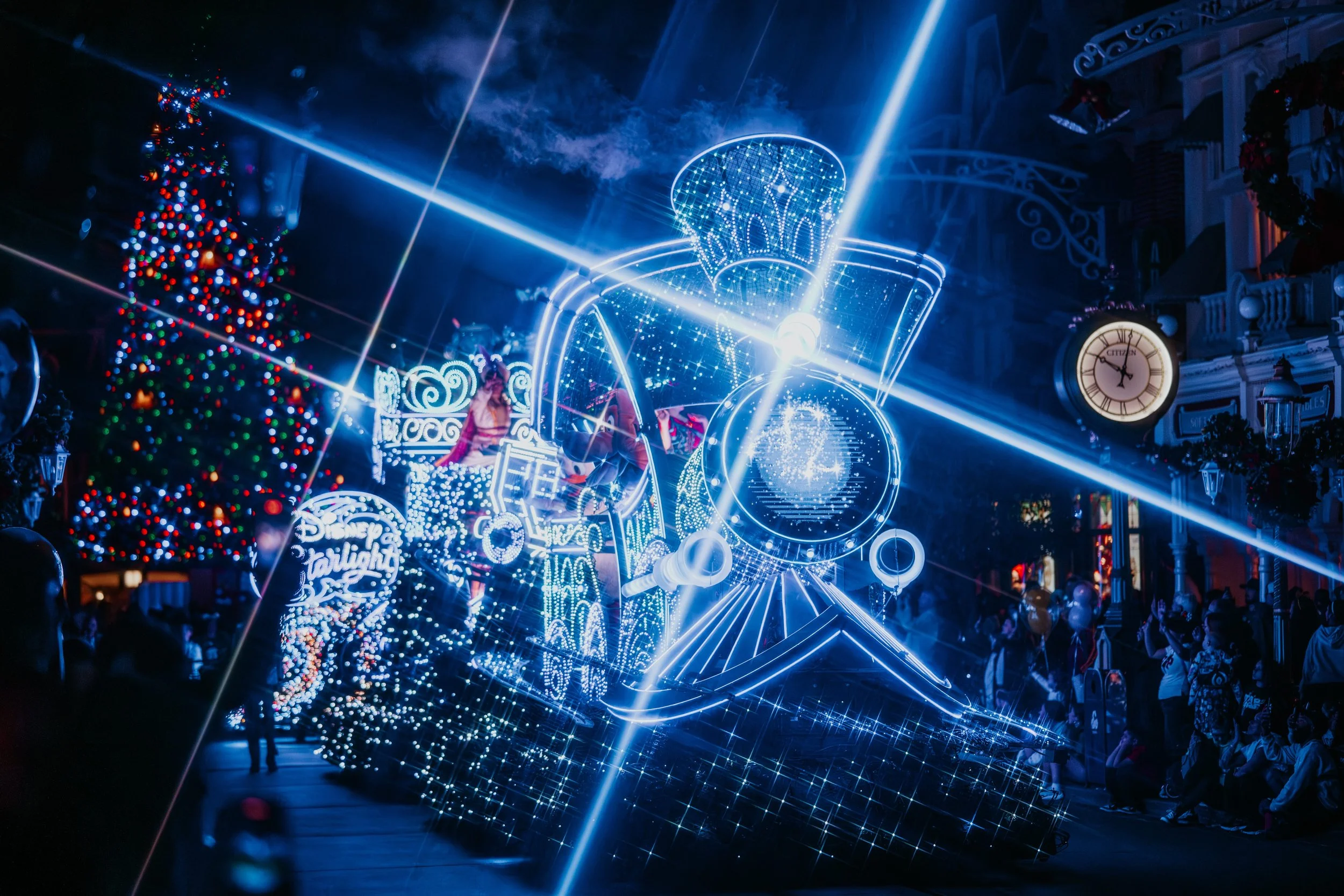 A brightly lit, illuminated float shaped like a train in a holiday parade, with Christmas decorations and a decorated Christmas tree in the background.