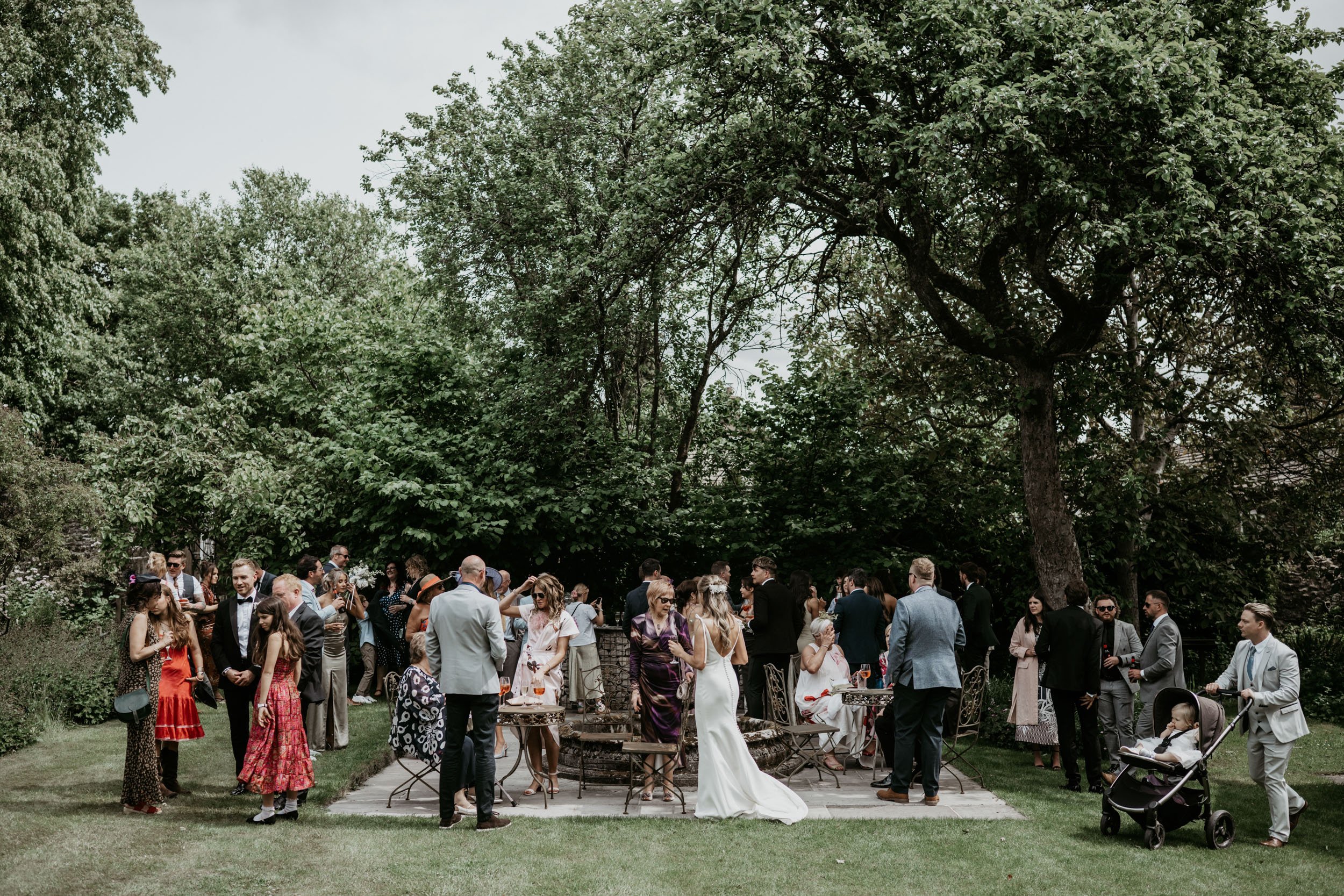 Guests mingling at an outdoor wedding reception in a lush green garden with a large tree in the background. Some guests are dressed in formal attire, including a woman in a white wedding dress, while others are in semi-formal and casual wear. A child is seated in a stroller on the right side.