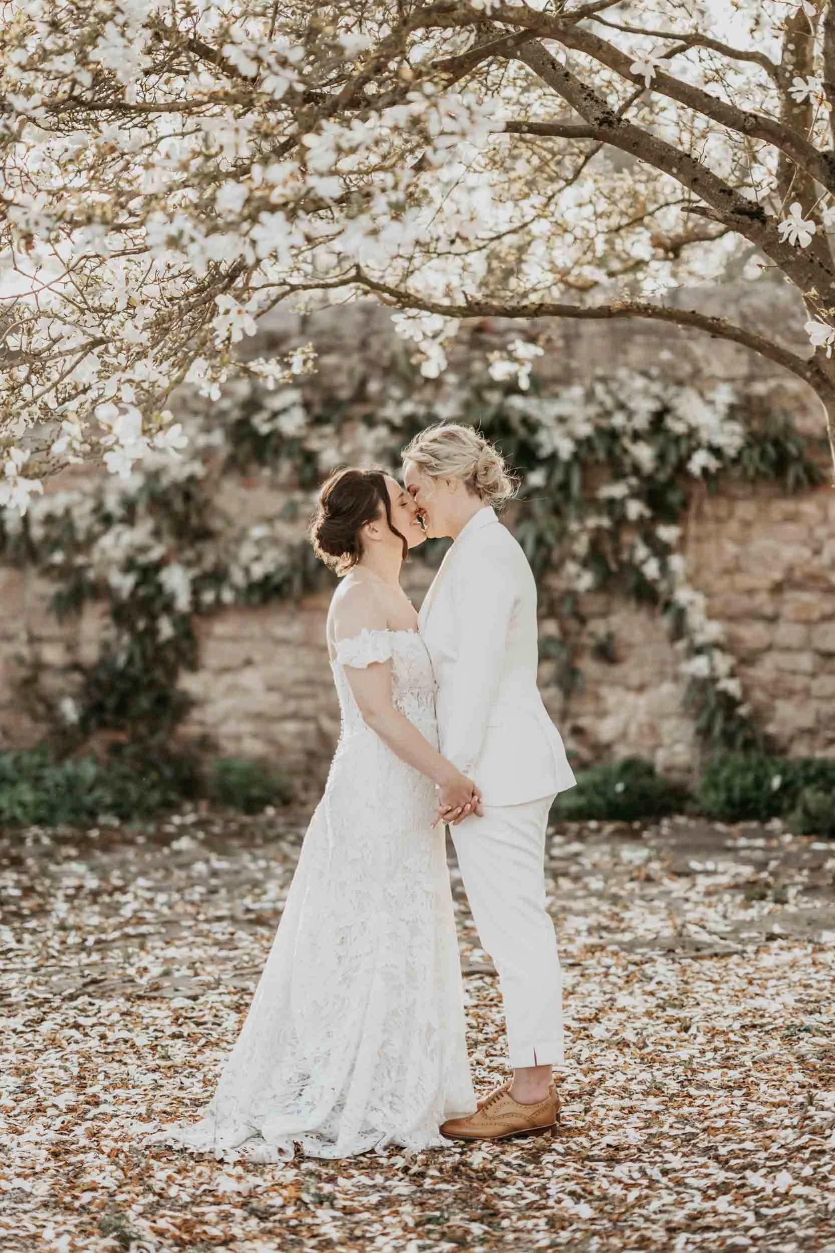 Two women, one in a wedding dress and the other in a white suit, stand close together under blossoming trees, holding hands and touching foreheads, in a romantic outdoor setting.