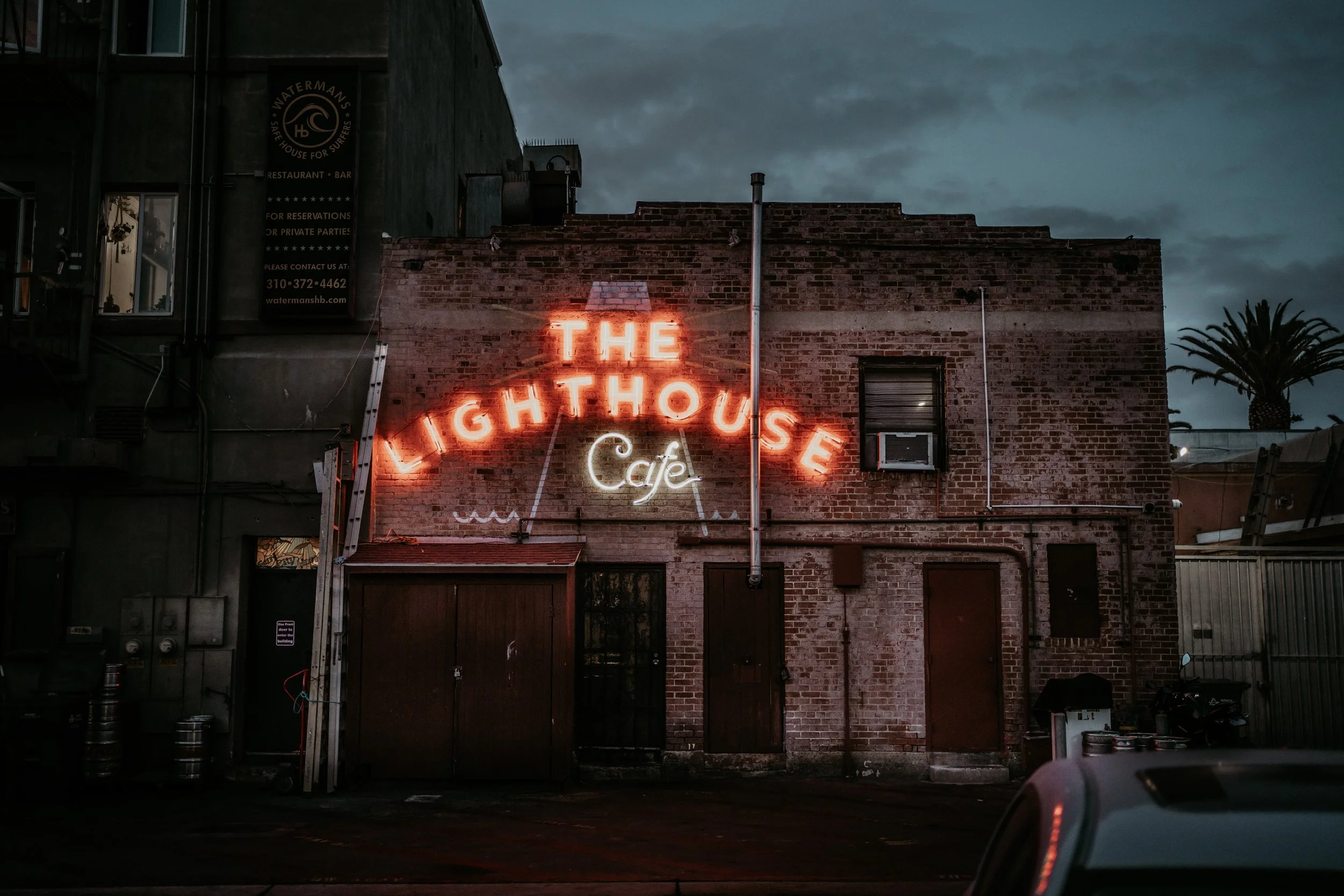 Neon sign on a brick building reading 'The Lighthouse Cafe' at night with cloudy sky and palm tree in the background.