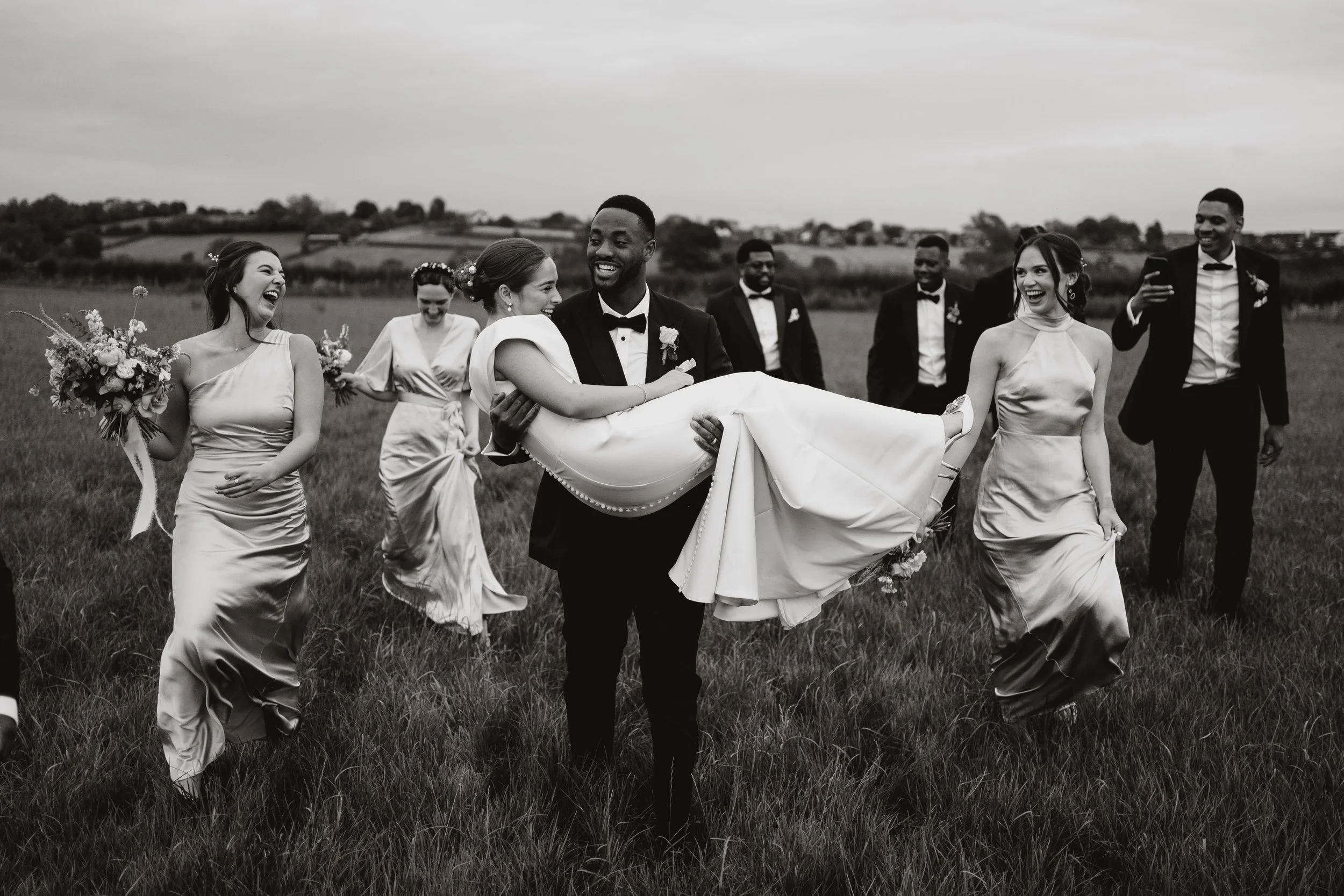 A wedding party in a field, with groom carrying bride, surrounded by bridesmaids and groomsmen, all smiling and celebrating outdoors.