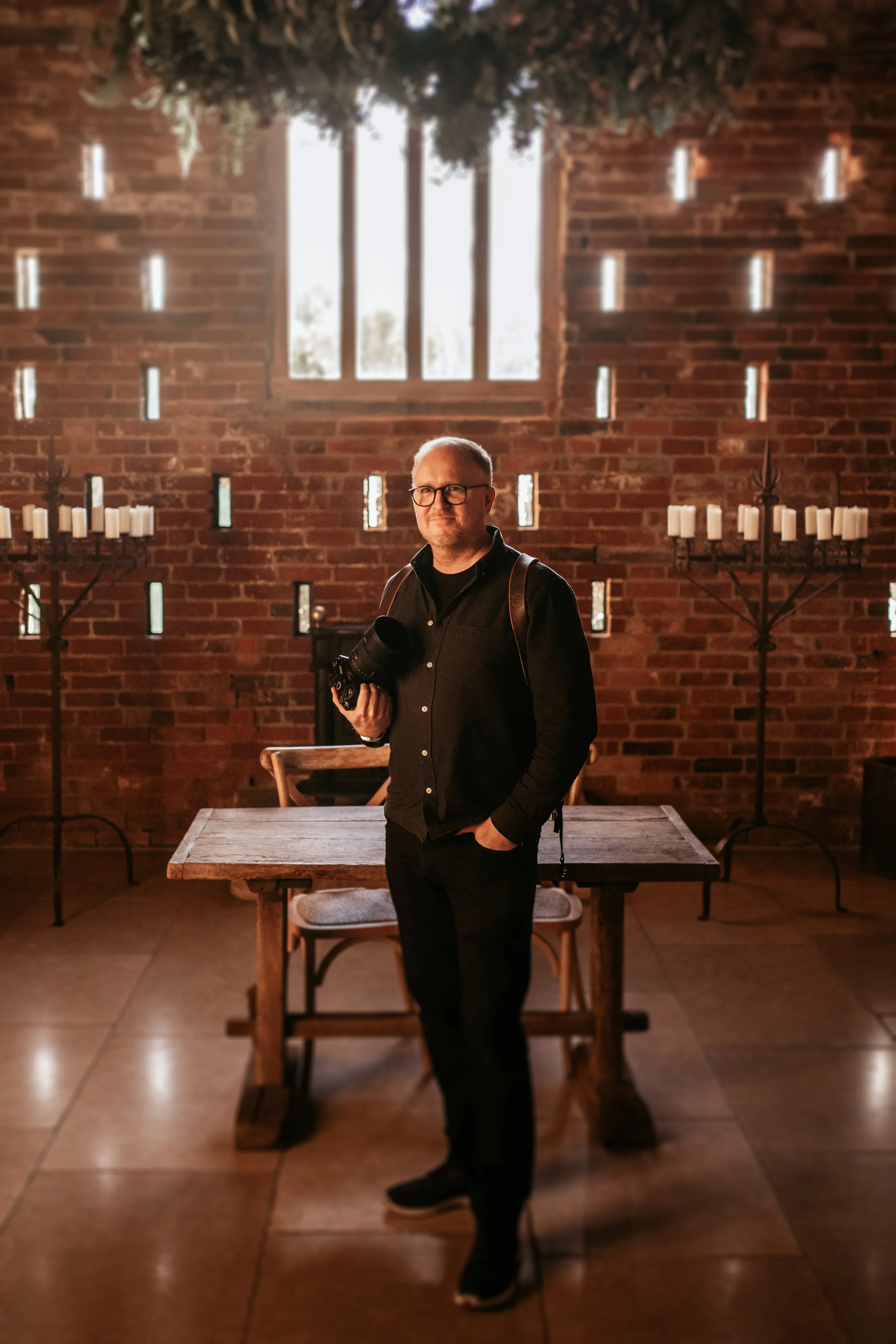 A man with glasses and a camera stands in a rustic brick room with candles and a large window.