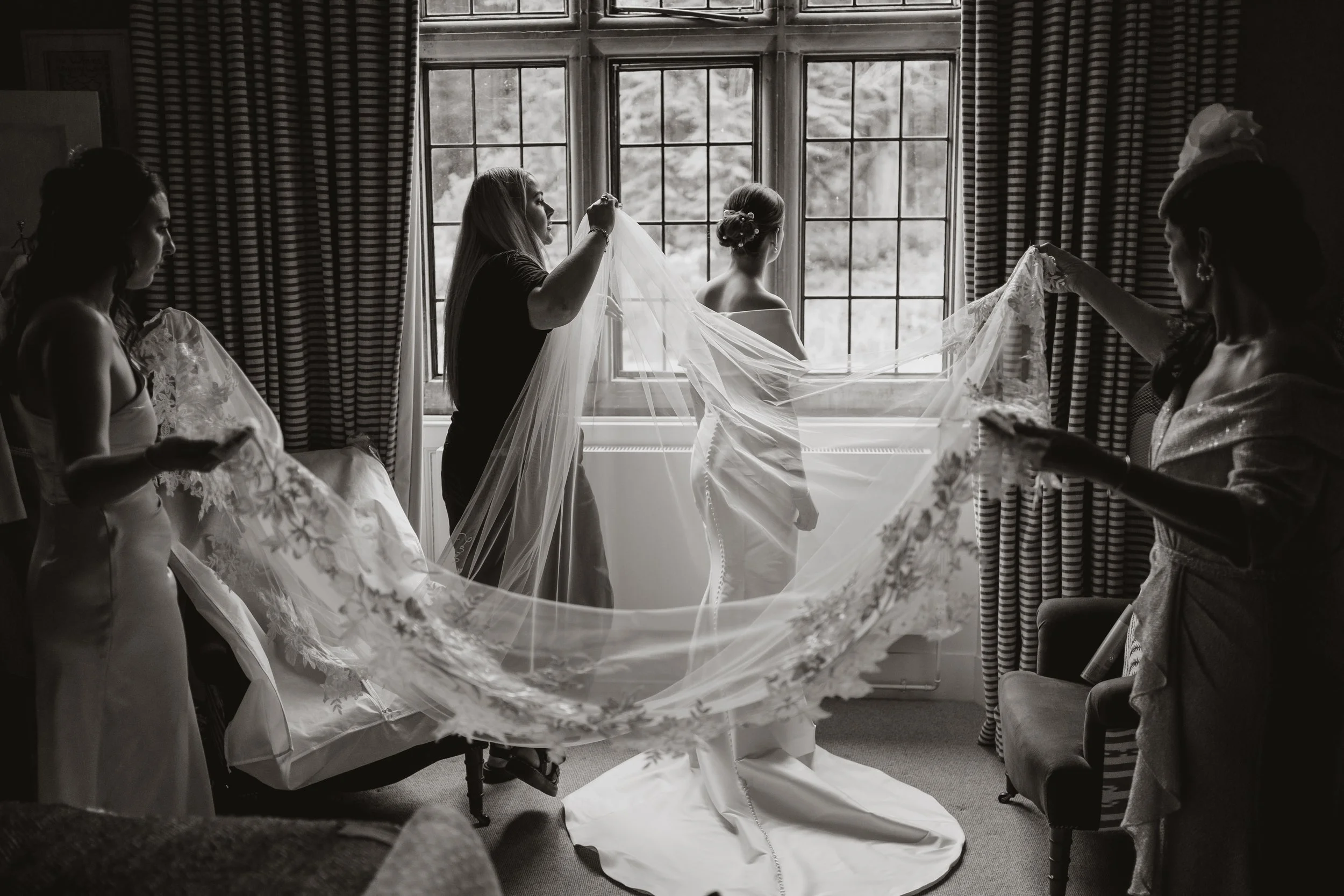 A bride stands by a large window with her wedding veil being arranged by four women, two of whom are holding the veil while the bride stands in a wedding dress. The scene is in a room with striped curtains and natural light coming through the window.