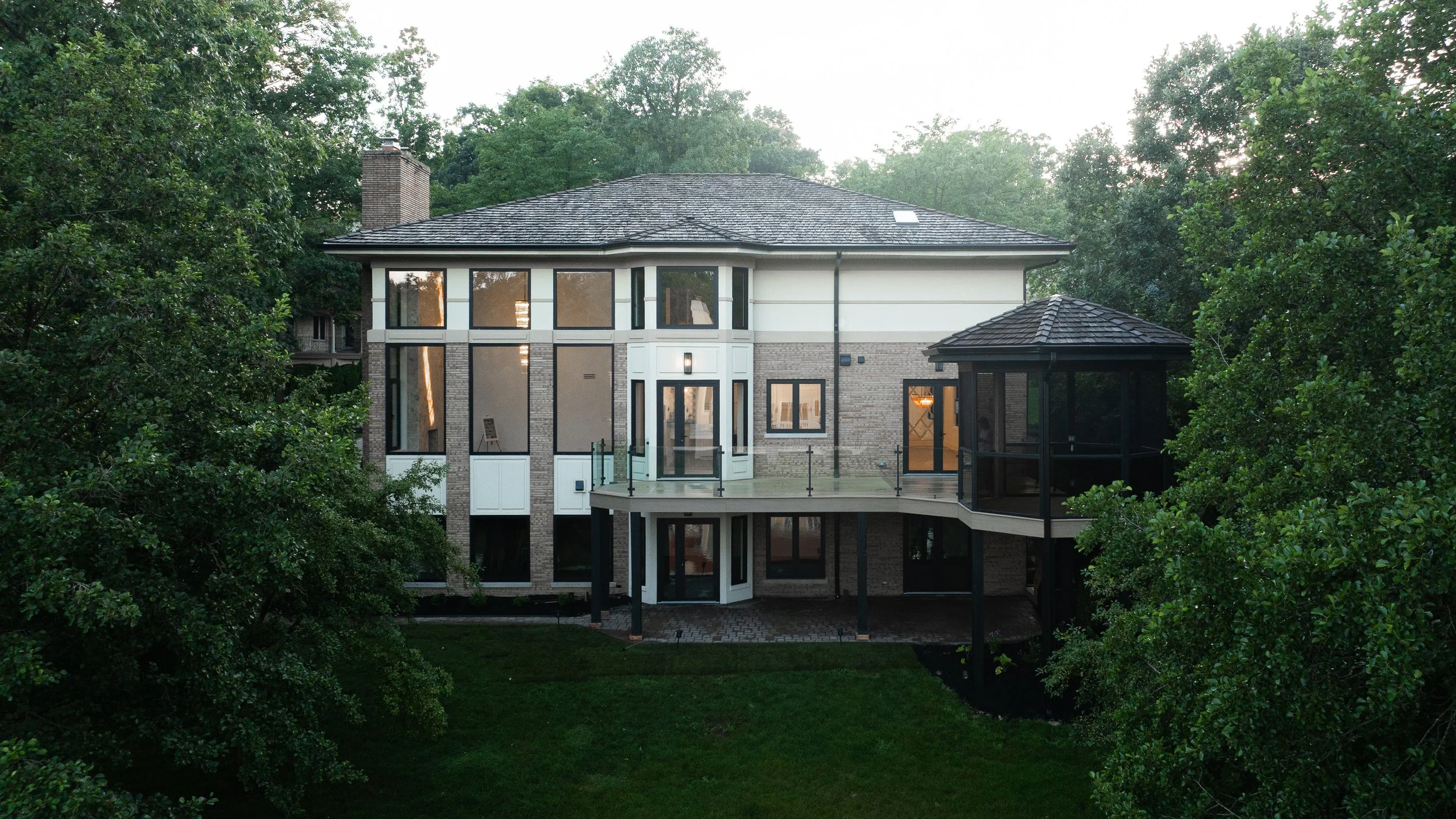 A large modern house with multiple stories, large windows, and a balcony, surrounded by trees, in a lush green environment.