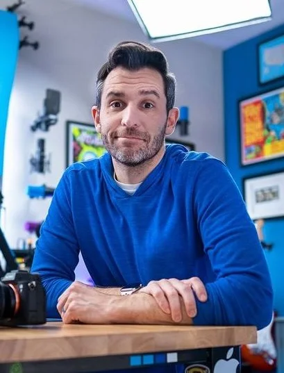 Tom Buck wearing a blue sweater sitting at a desk in a tech store or studio with monitors and colorful artwork in the background.
