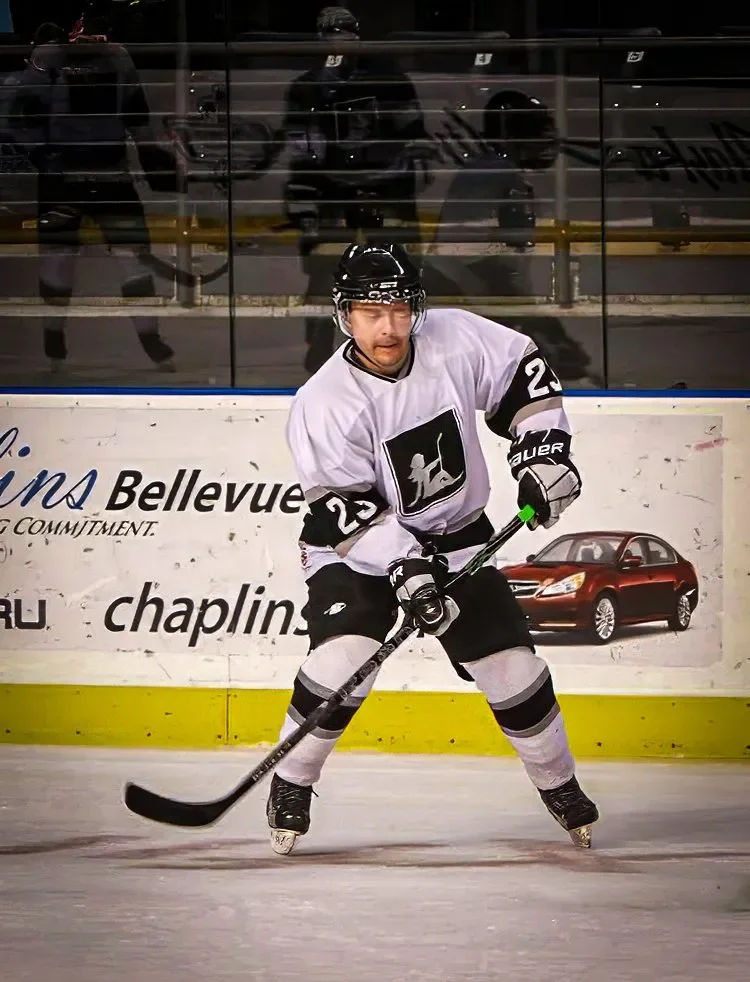 Joel Harris in a white jersey with black and grey gear, skating on the ice with a hockey stick, behind a glass barrier in an indoor rink.