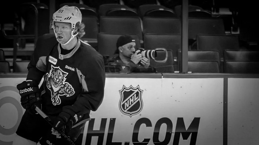 An ice hockey player in a black jersey with a Florida Panthers logo, wearing a helmet and gloves, standing on the ice rink. Behind the glass, a Joel Harris with a large camera lens is capturing the game.