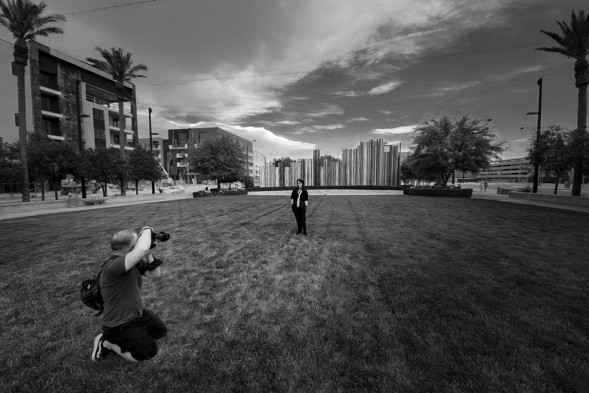Joel Harris kneeling on the grass with a camera, photographing a woman standing in the middle of the grass field in an urban park setting during daytime, with modern buildings, trees, streetlights, and a cloudy sky in the background.