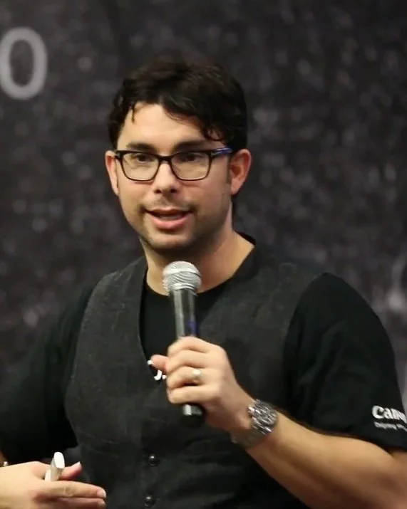 Roberto Valenzuela, glasses, wearing a black t-shirt and gray vest, holding a microphone, speaking at an event.