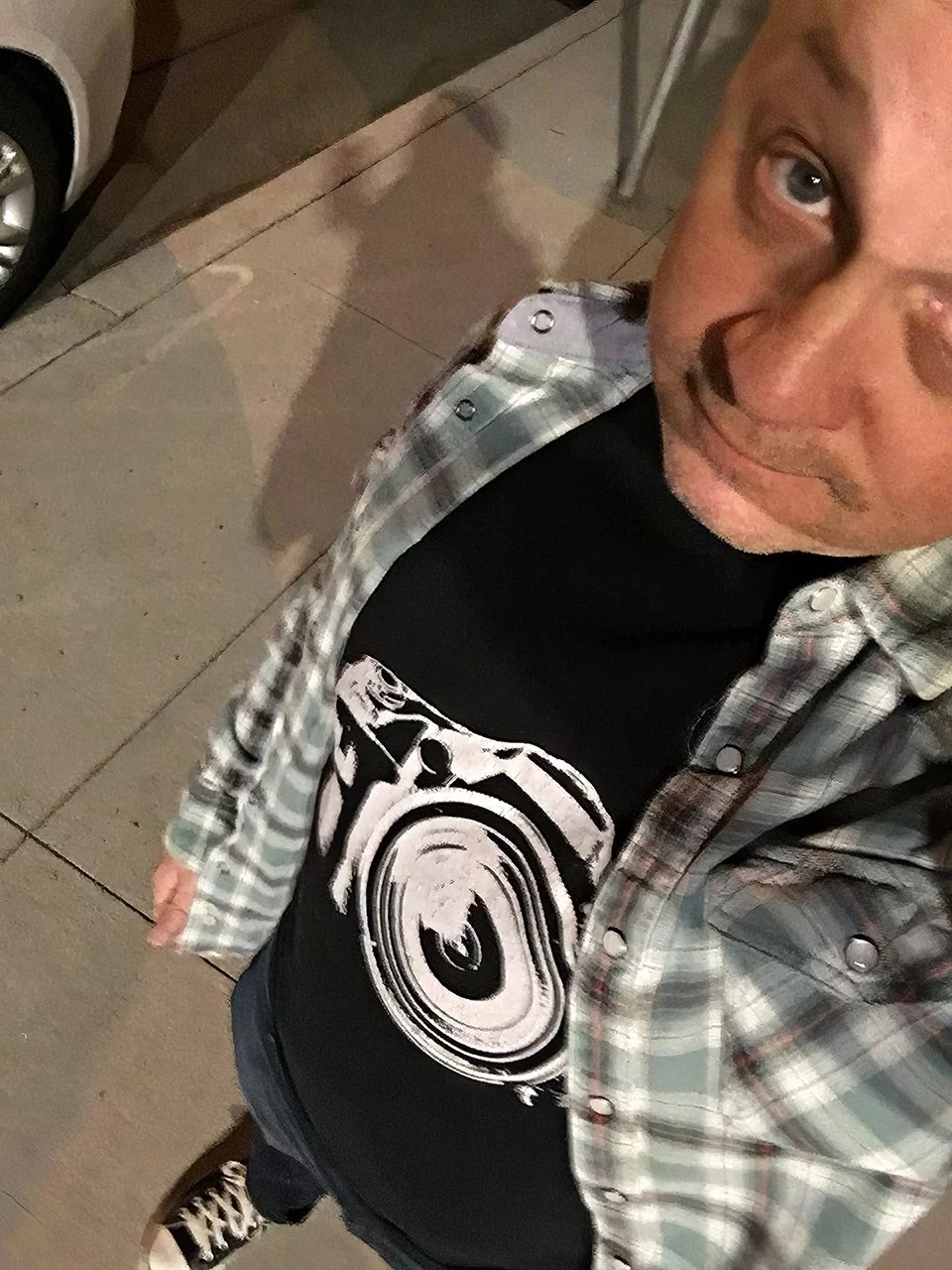 Joel Harris, taking a selfie from a top-down angle on a sidewalk at night. He is wearing a black T-shirt with a large white camera graphic and a plaid button-up shirt over it. The background shows part of a parked car and concrete sidewalk.