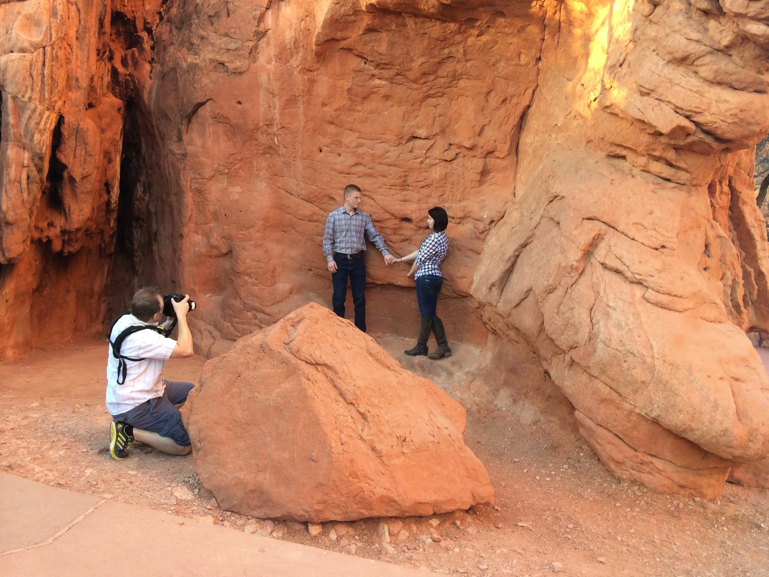 An engagement photo sesssion is taking place inside a large red sandstone canyon with three rocks. A man and a woman are holding hands, facing each other, with Joel Harris kneeling, taking their photo.