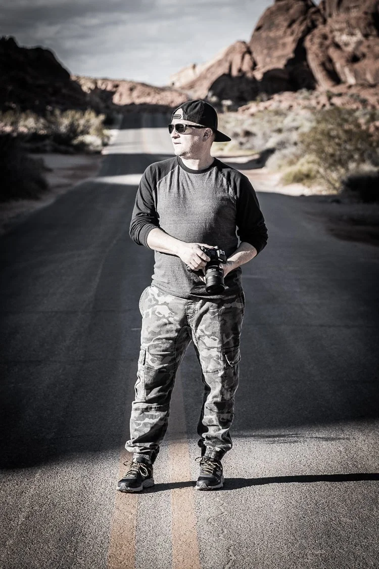 Joel Harris, from The Creative Urge, standing in the middle of a deserted road in Valley of Fire State Park, holding a camera, wearing sunglasses, a backwards cap, a long-sleeved shirt, camouflage pants, and hiking shoes.