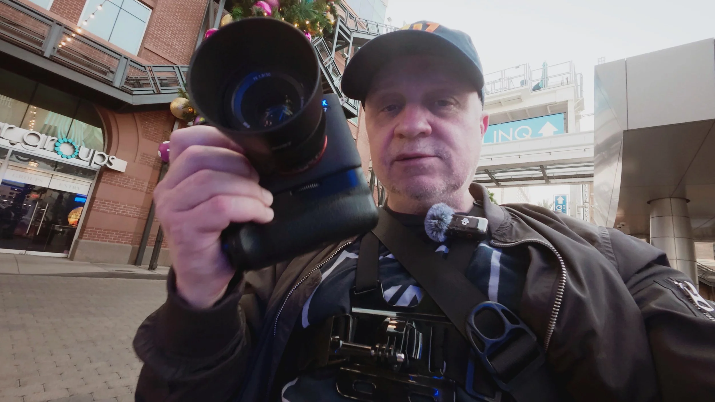 Joel Harris in a video from The Creative Urge, holding a camera, standing outside in an urban area with a brick building in the background.