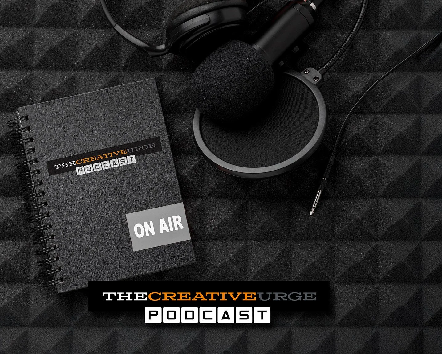 A black podcast recording setup including a notebook labeled 'The Creative Urge Podcast' with an 'On Air' sticker, a microphone with a foam windscreen, headphones, and an audio jack on a black soundproof foam surface.