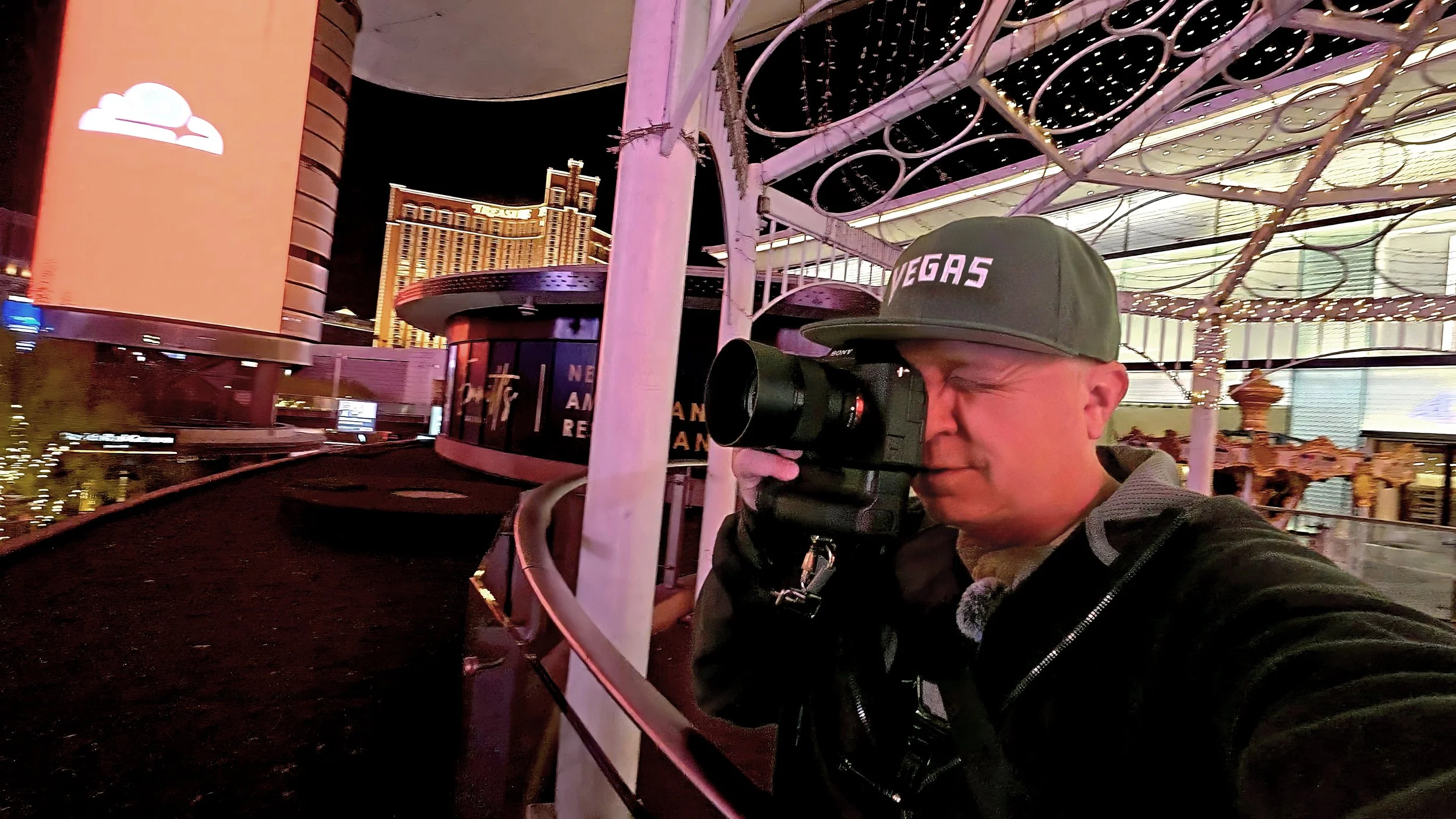 Joel Harris in a video from The Creative Urge, holding a camera, standing outside in an urban area with a metal sculpture in the background.