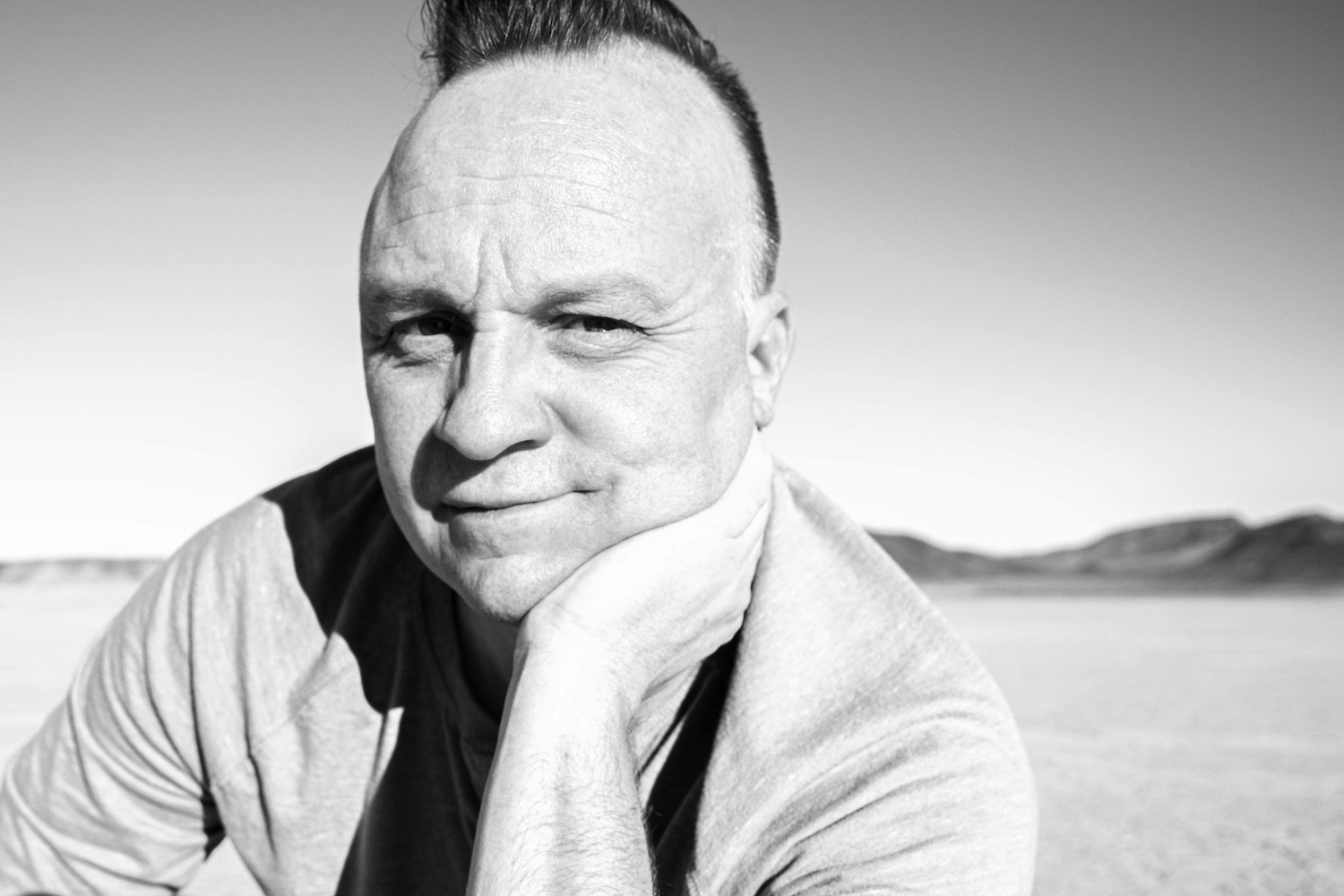A black and white photo of Joel Harris, founder of The Creative Urge, resting his chin on his hand, outdoors with a flat landscape and hills in the background.