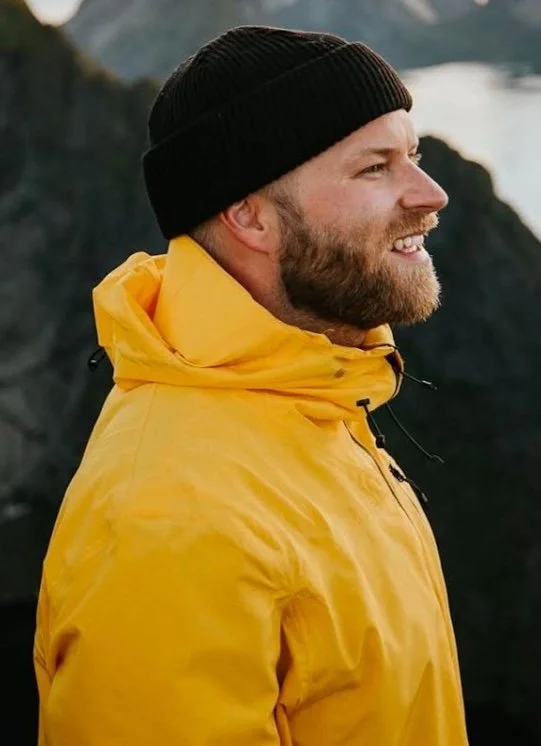 Teppo Haapoja in a yellow jacket and black beanie outdoors near water and mountains, smiling.