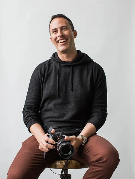 A smiling man sitting on a stool holding a camera, against a plain light gray background.